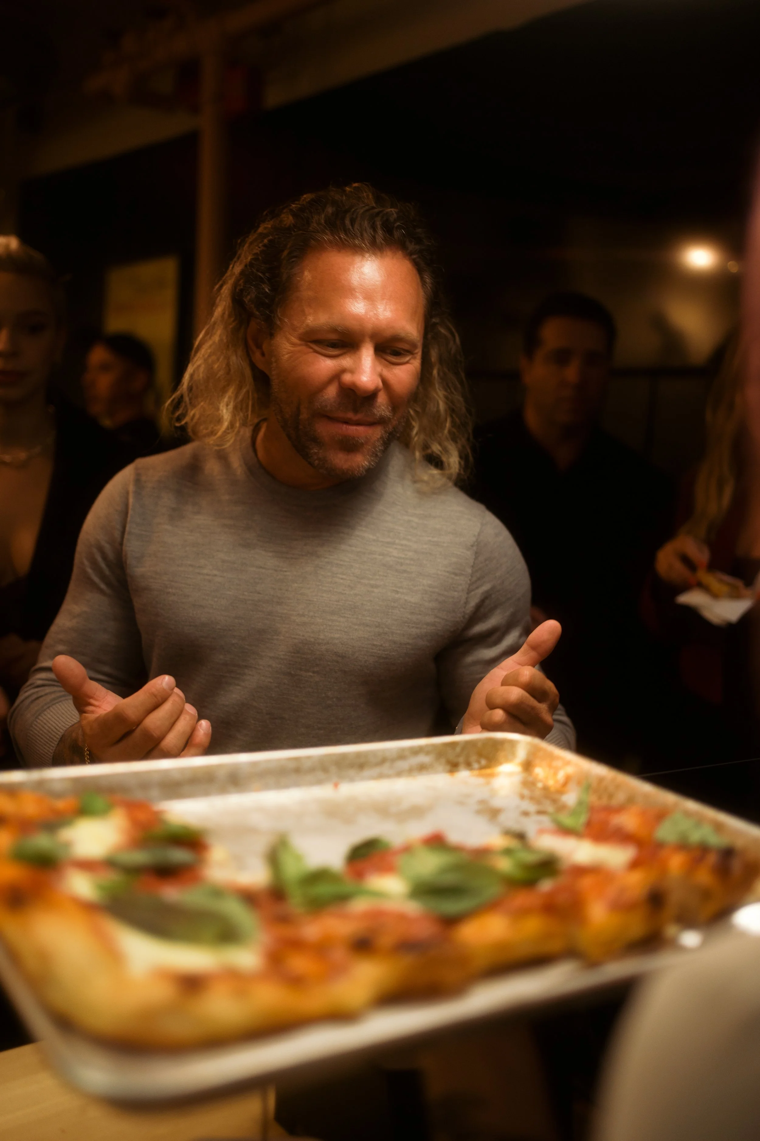 A man with long hair and a gray shirt looking at a pizza with basil leaves, surrounded by people in a dimly lit room.