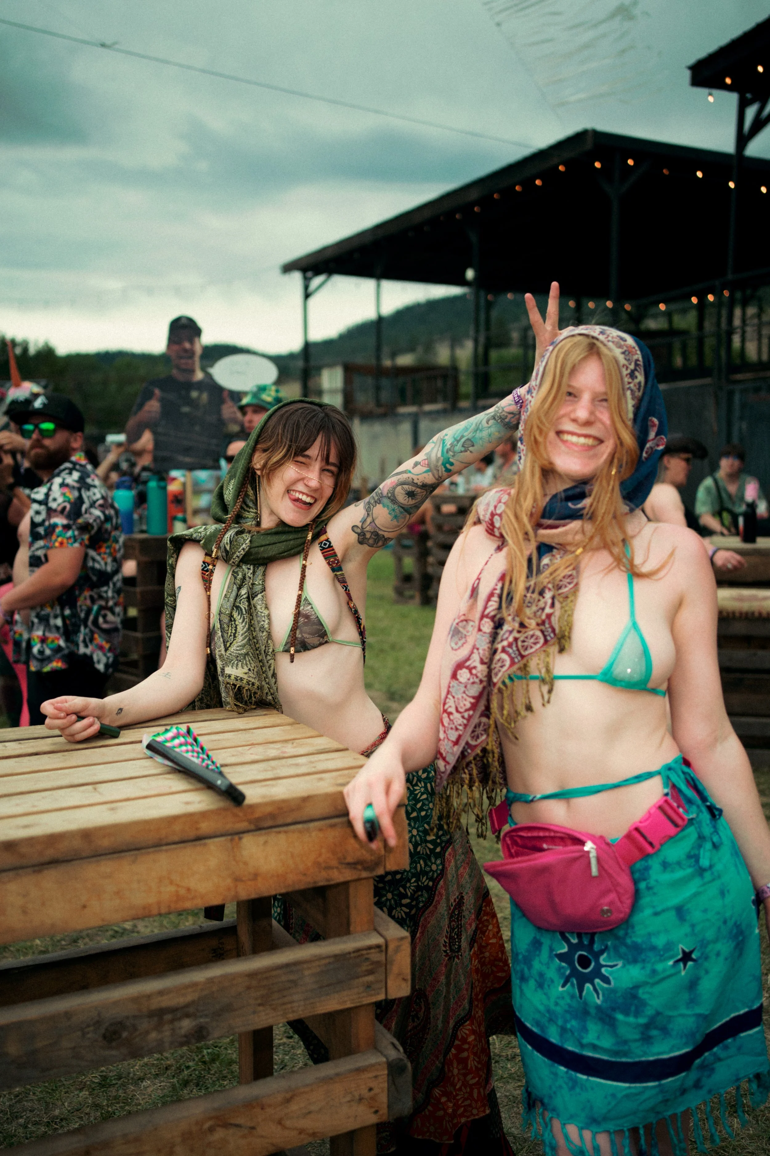 Two young women smiling and having fun at an outdoor event, with one giving the peace sign behind the other, both dressed in colorful, minimal clothing and headscarves, during daytime with a cloudy sky.
