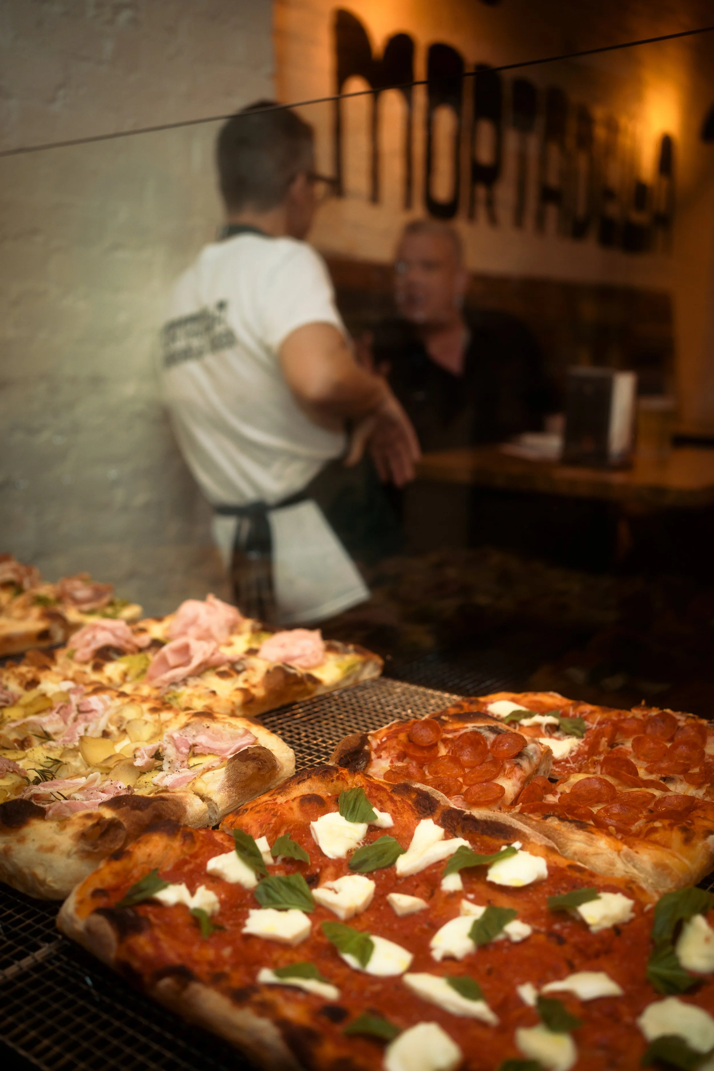 Assorted pizzas with toppings like pepperoni, cheese, and ham displayed in a pizzeria, with two people visible in the background behind a glass window.
