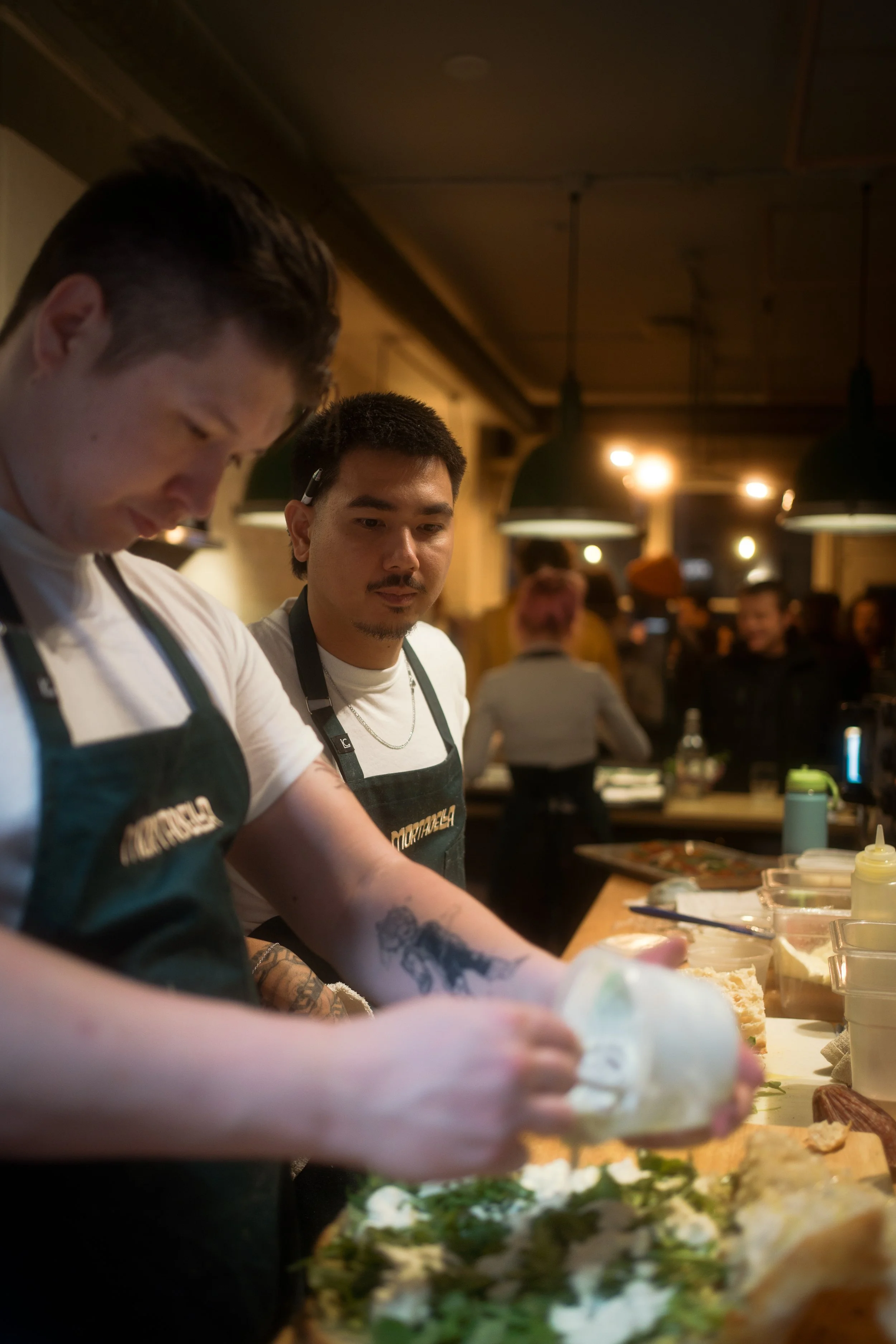 Two men wearing aprons preparing food in a restaurant kitchen with people and hanging lights in the background.