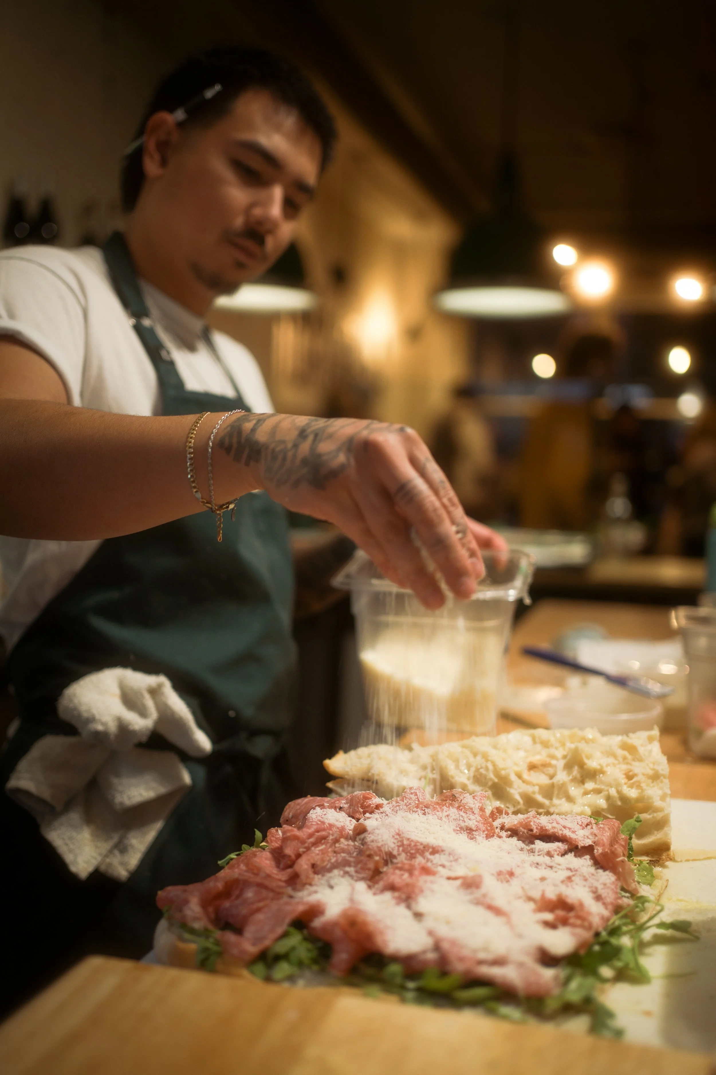 A man preparing food in a restaurant kitchen, seasoned meat and ingredients on a cutting board, with warm lighting and background customers.