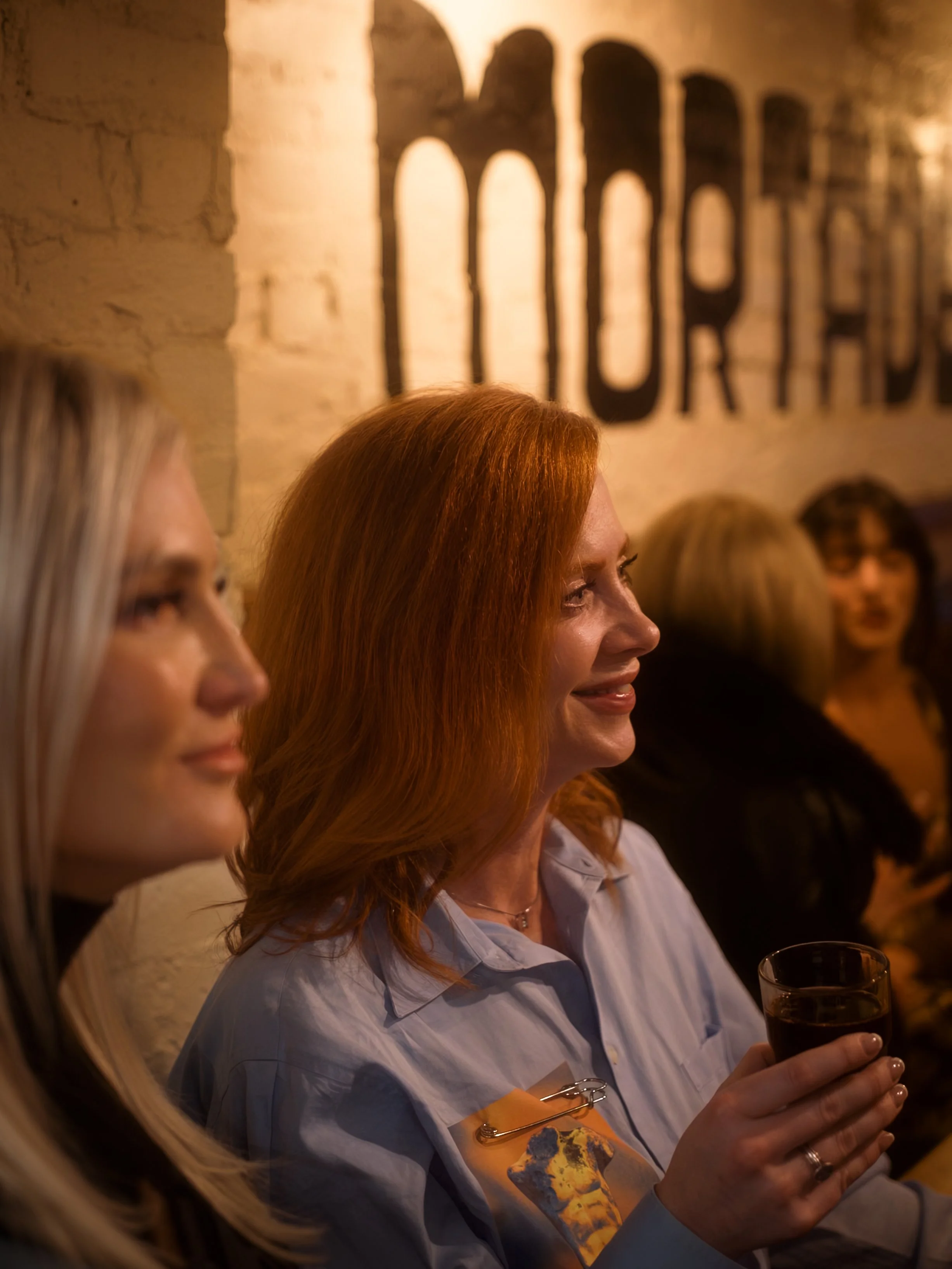 A woman with red hair holding a glass of dark beverage, surrounded by other people, in a dimly lit, rustic indoor setting with a wall featuring large black text.