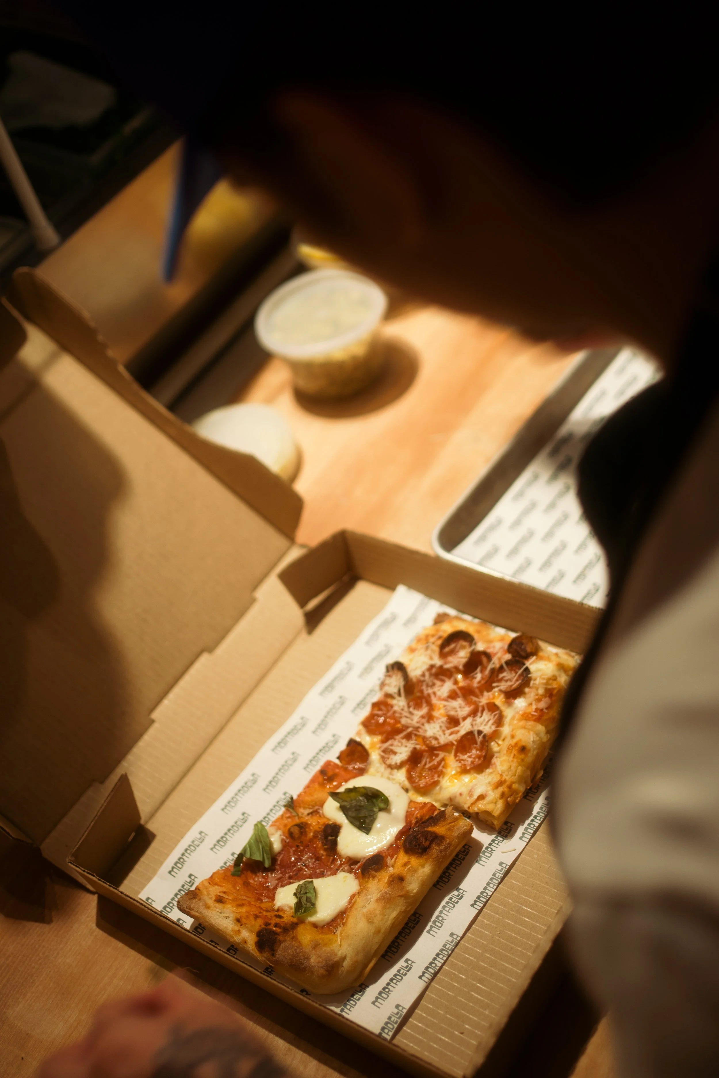 Open pizza box containing two slices of pizza, one with pepperoni and the other with cheese and basil.