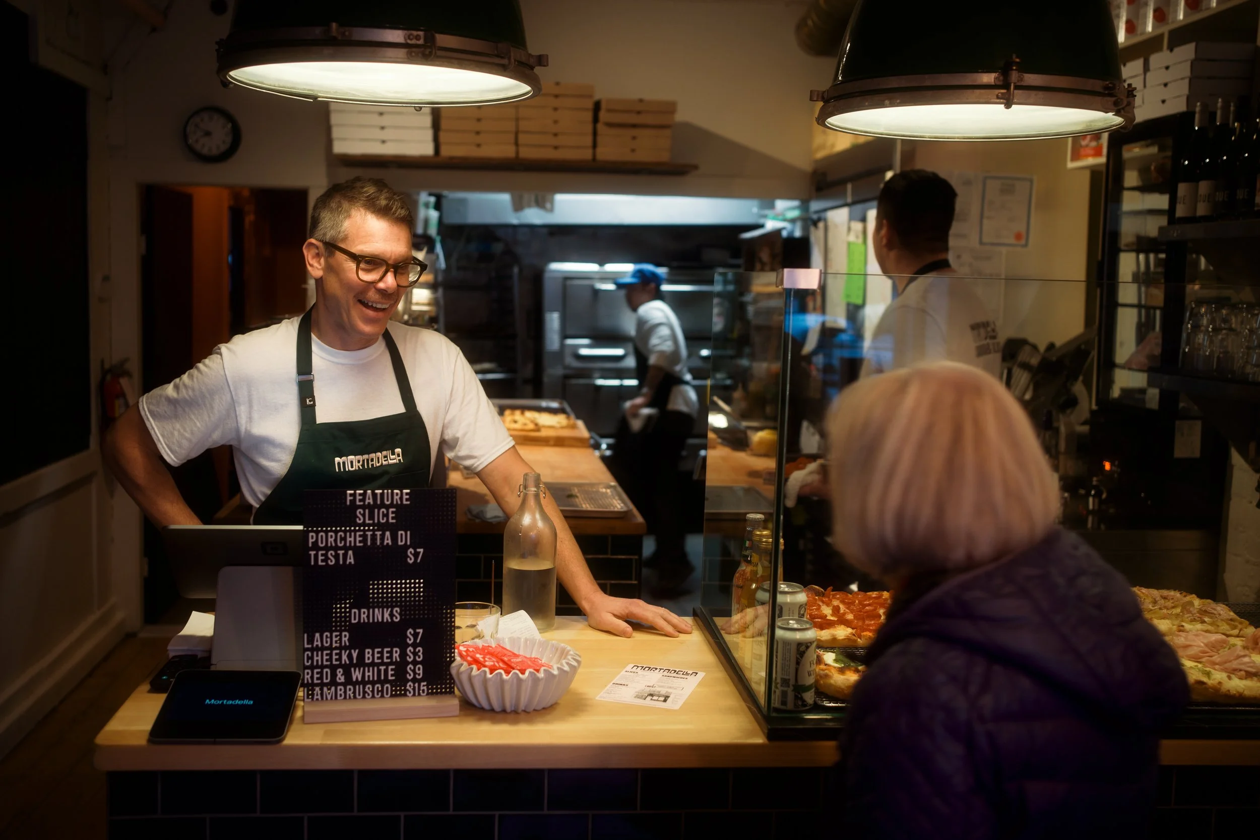 A smiling man with glasses wearing a white t-shirt and black apron serving a customer at a pizza shop counter, with the kitchen in the background.