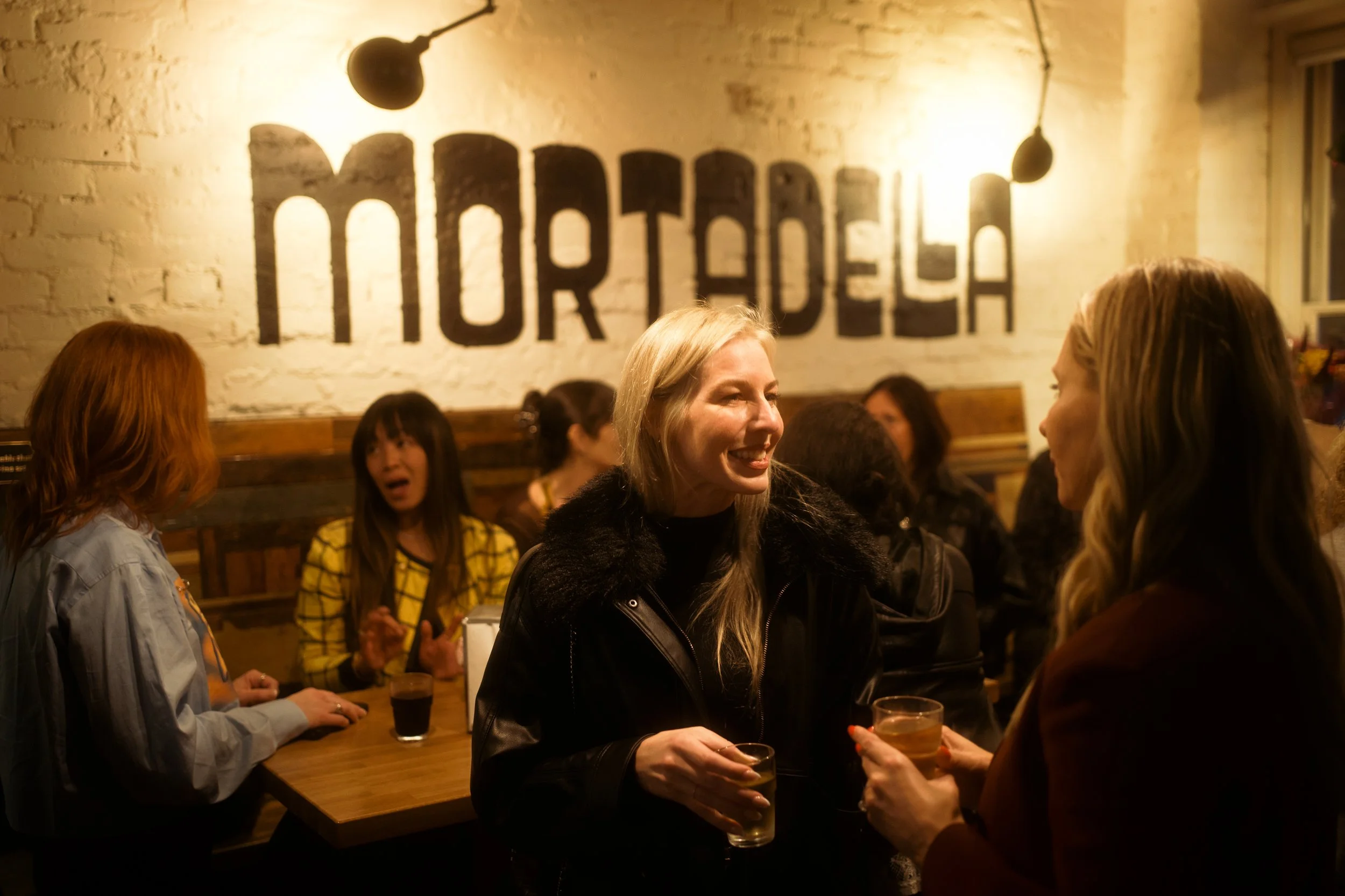 People socializing at a bar or restaurant with a large 'MORTADEL