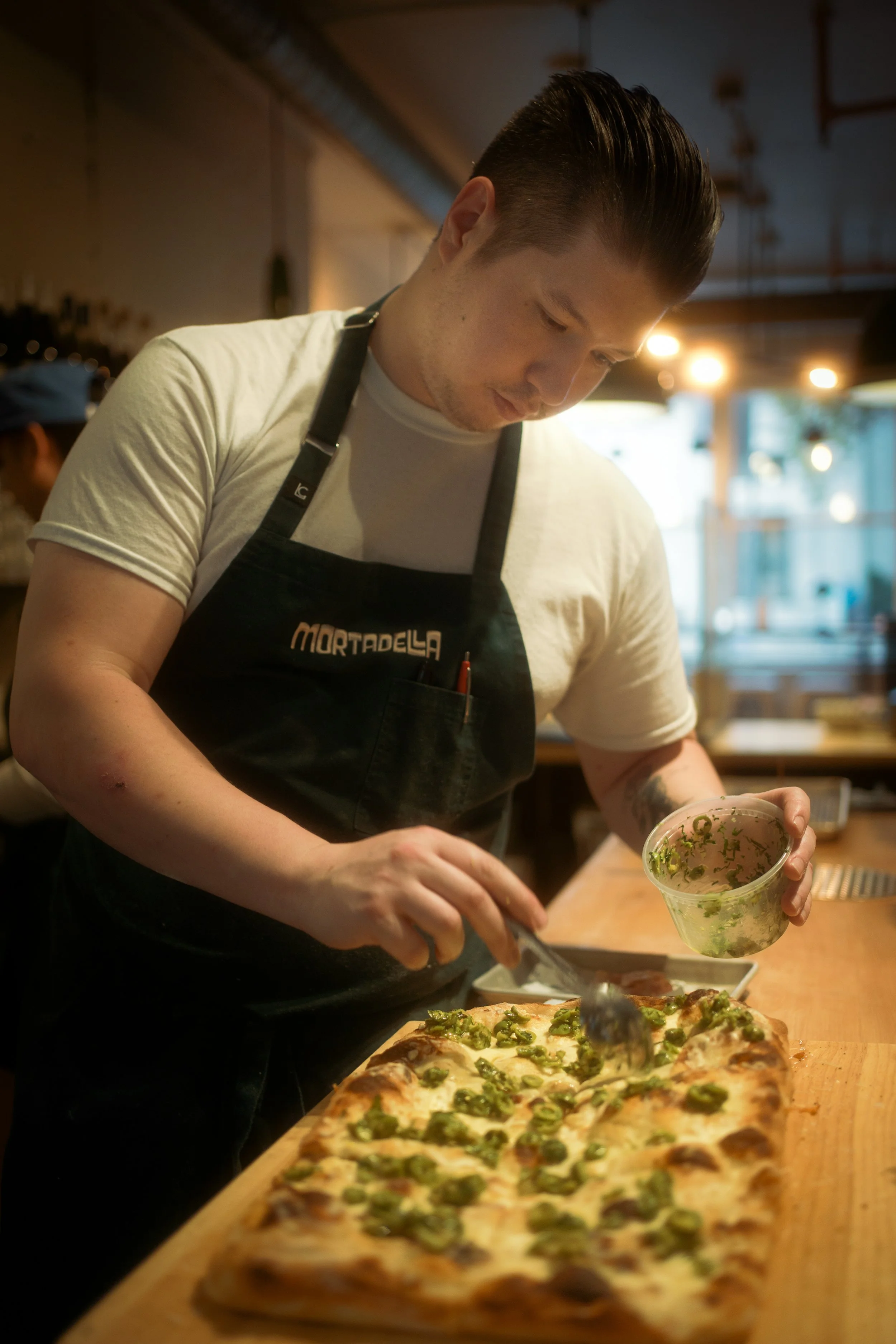 A chef is preparing a rectangular pizza topped with green herbs and slices of green peppers in a restaurant kitchen.