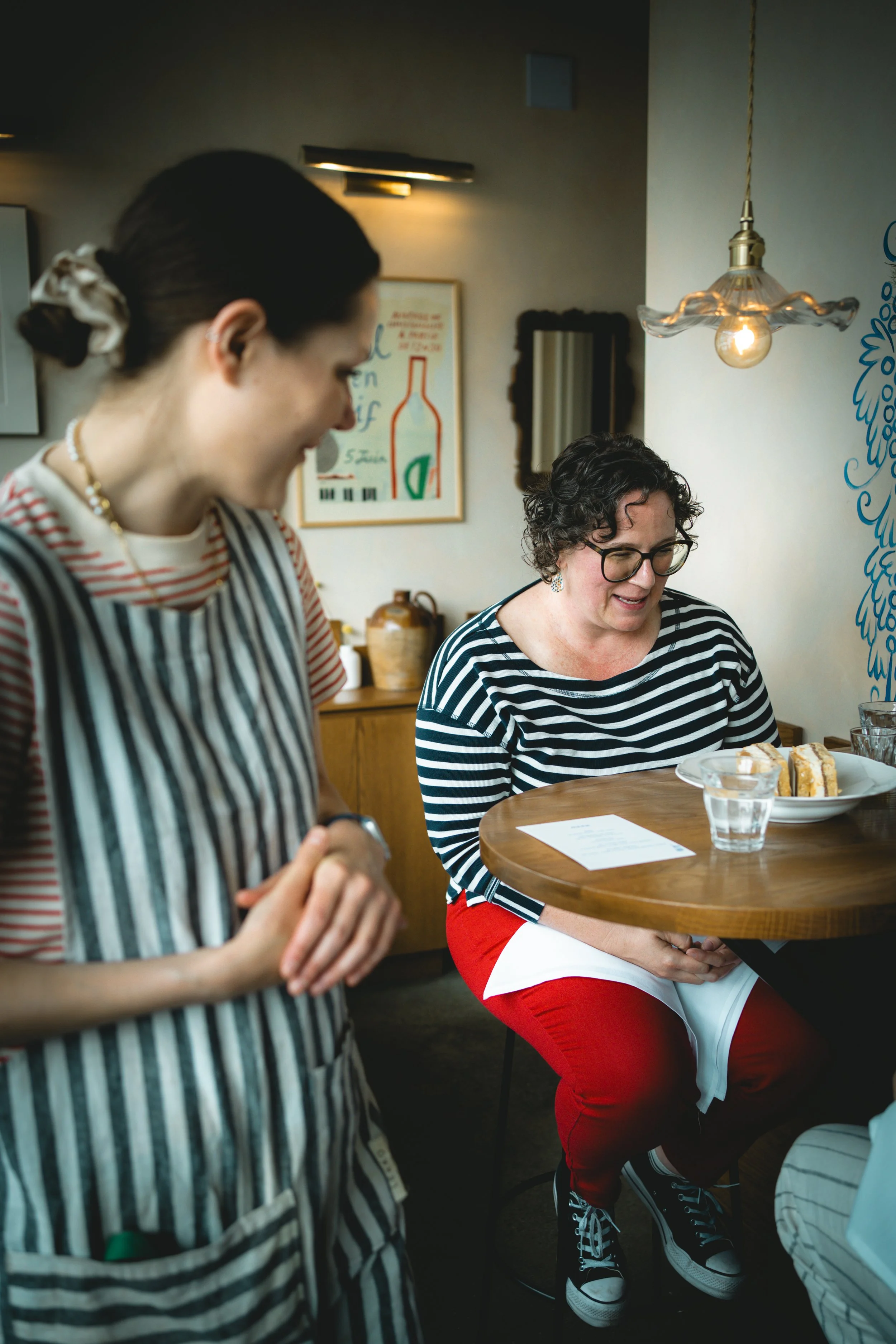 A woman in a striped shirt signing or reading a paper while seated at a wooden table with desserts, with a server in the foreground in a striped apron.