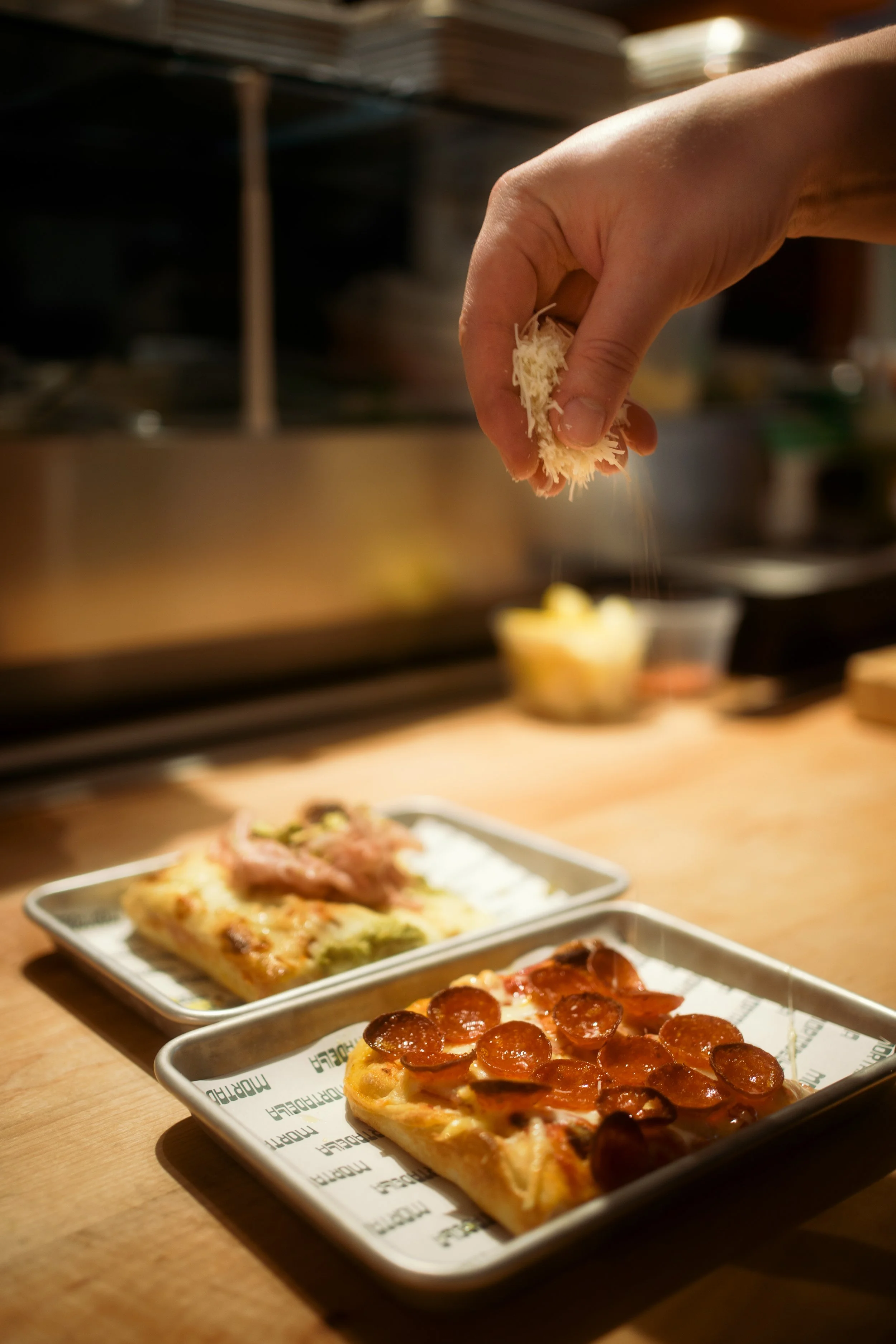 Hand sprinkling grated cheese on two slices of pizza, one topped with pepperoni and the other with ham and vegetables, on a wooden table in a restaurant or kitchen.
