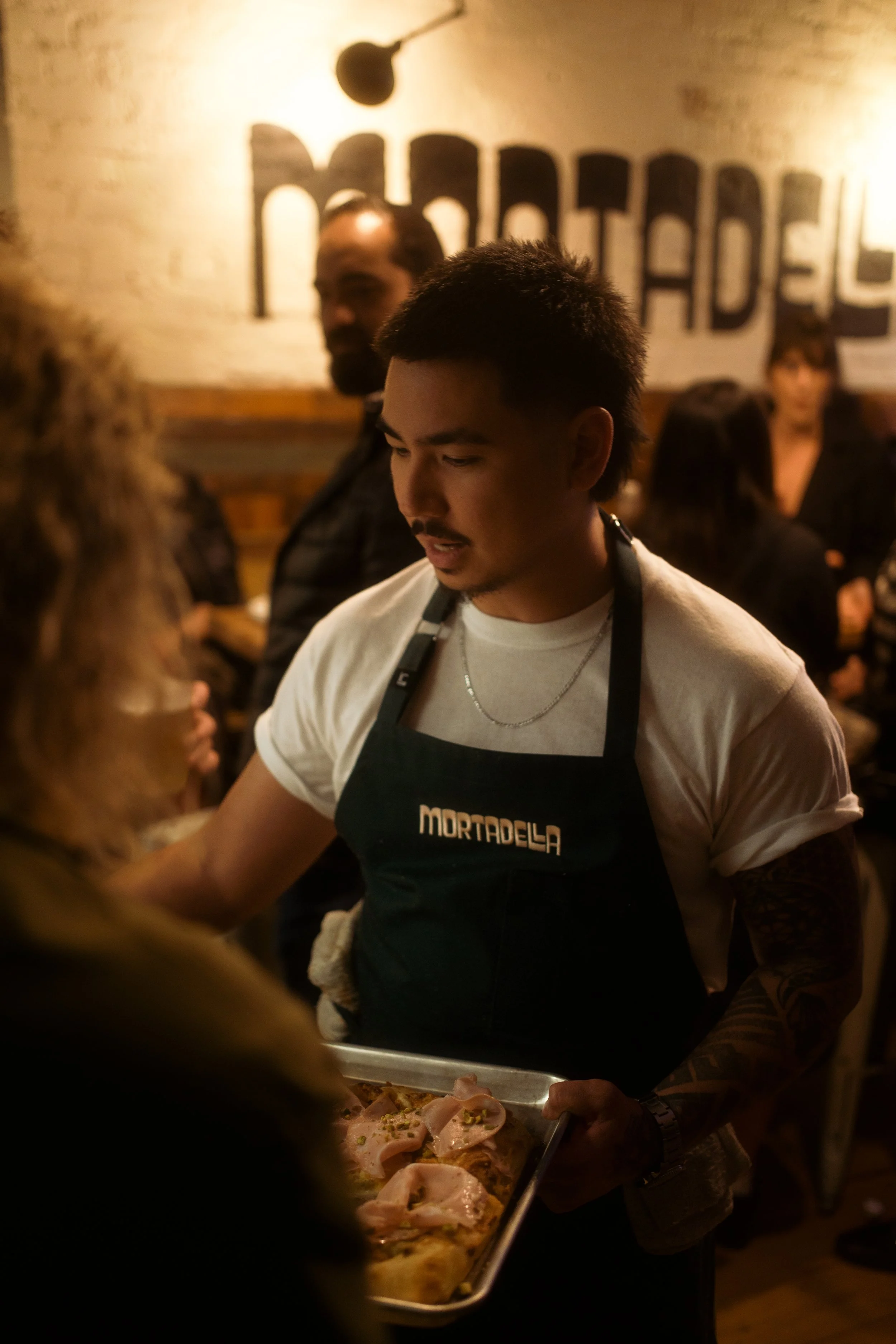 A man wearing a black apron with 'MORTADELA' on it, serving food at a restaurant, with a tray of raw chicken and meat. The background shows a brick wall with the word 'MORTADELA' and a hanging lamp.