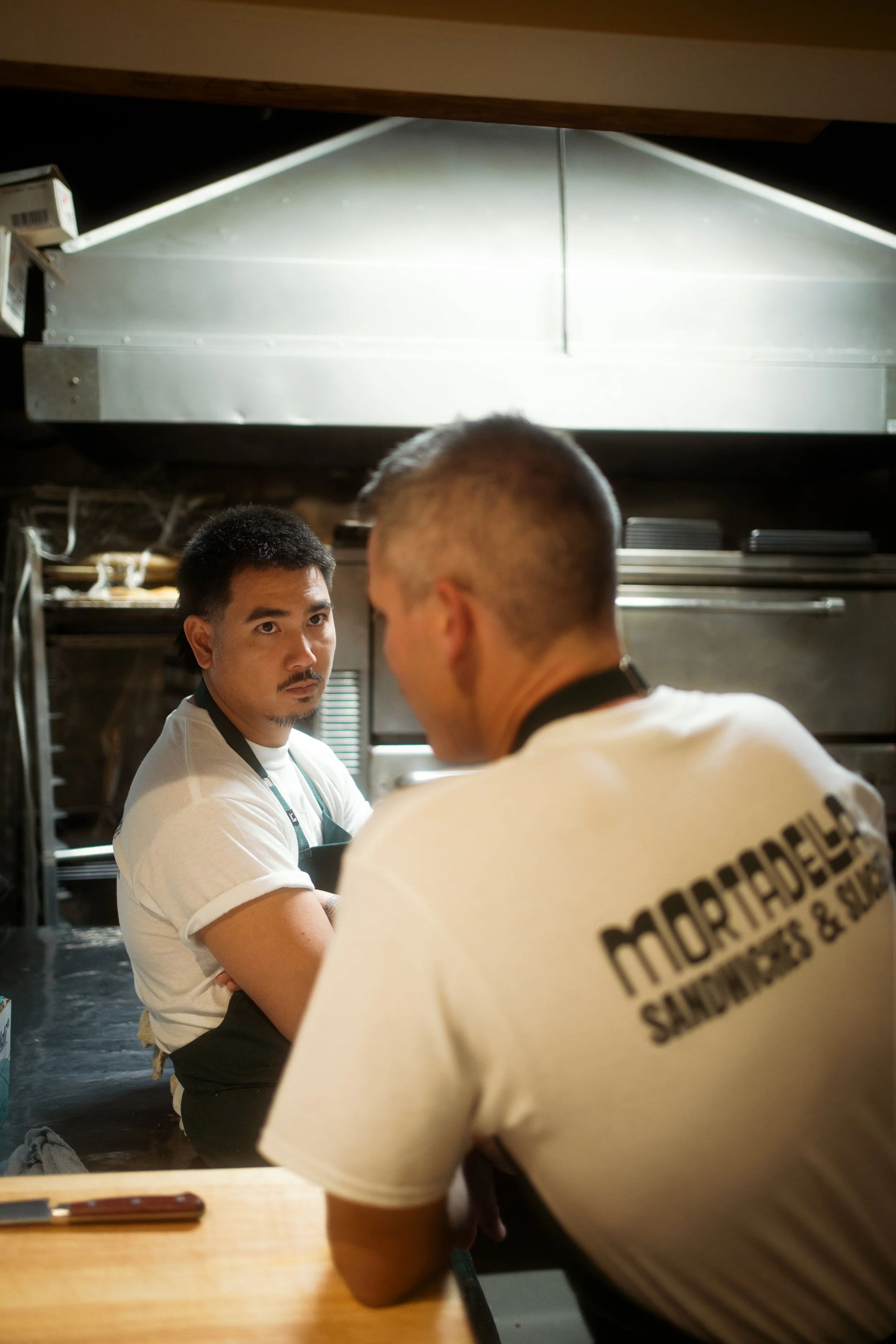 Two male chefs in a restaurant kitchen, one facing the camera with a serious expression and the other with his back turned, wearing a shirt that says 'Midoriya'.