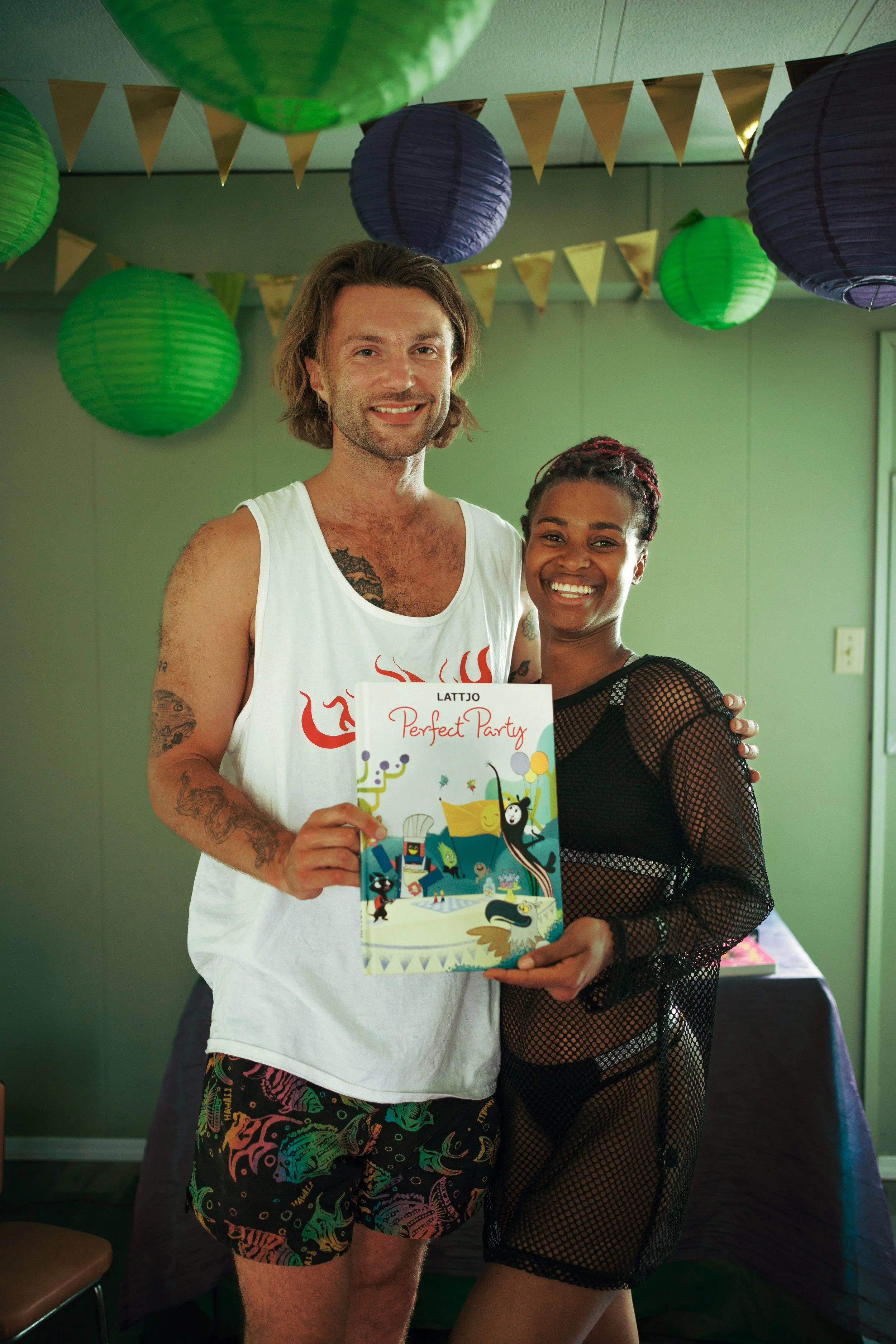 Two smiling people, a man and a woman, standing together at a party decorated with green and purple lanterns and gold pennant banners. They are holding a colorful party booklet that reads 'Perfect Party'.