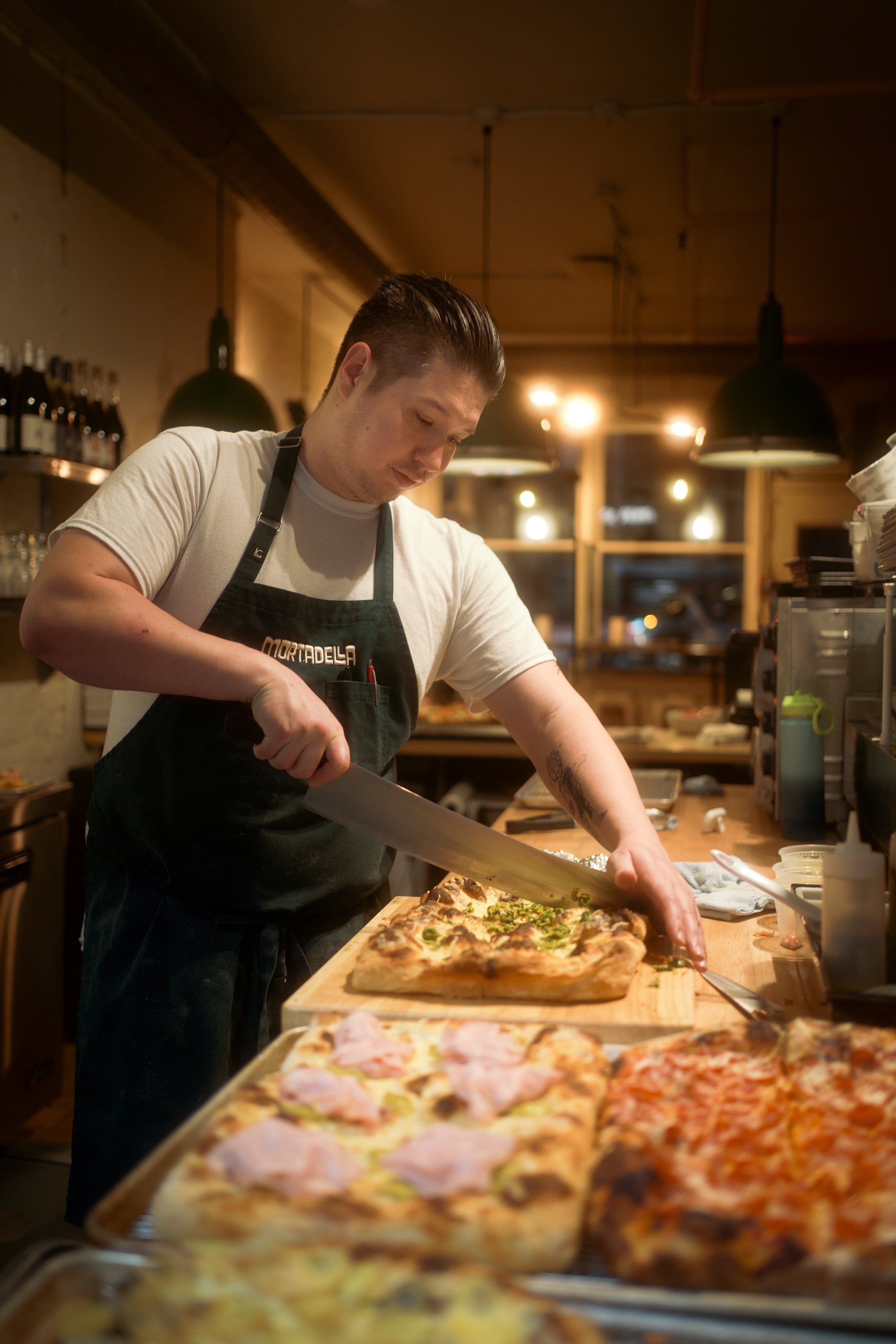A man wearing a black apron is slicing a pizza at a counter in a dimly lit restaurant kitchen. There are other pizzas in the foreground and bottles on a shelf in the background.