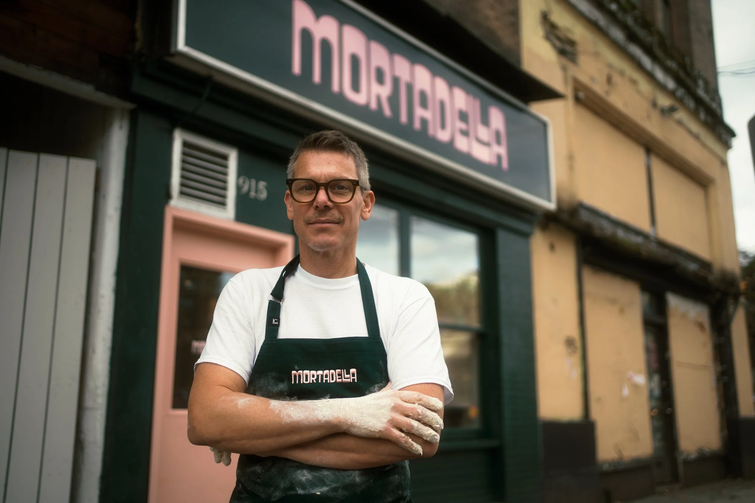 A man wearing glasses and a white shirt stands outside a bakery named Mortadela, with his arms crossed and flour on his hands, in front of the storefront with a neon sign.