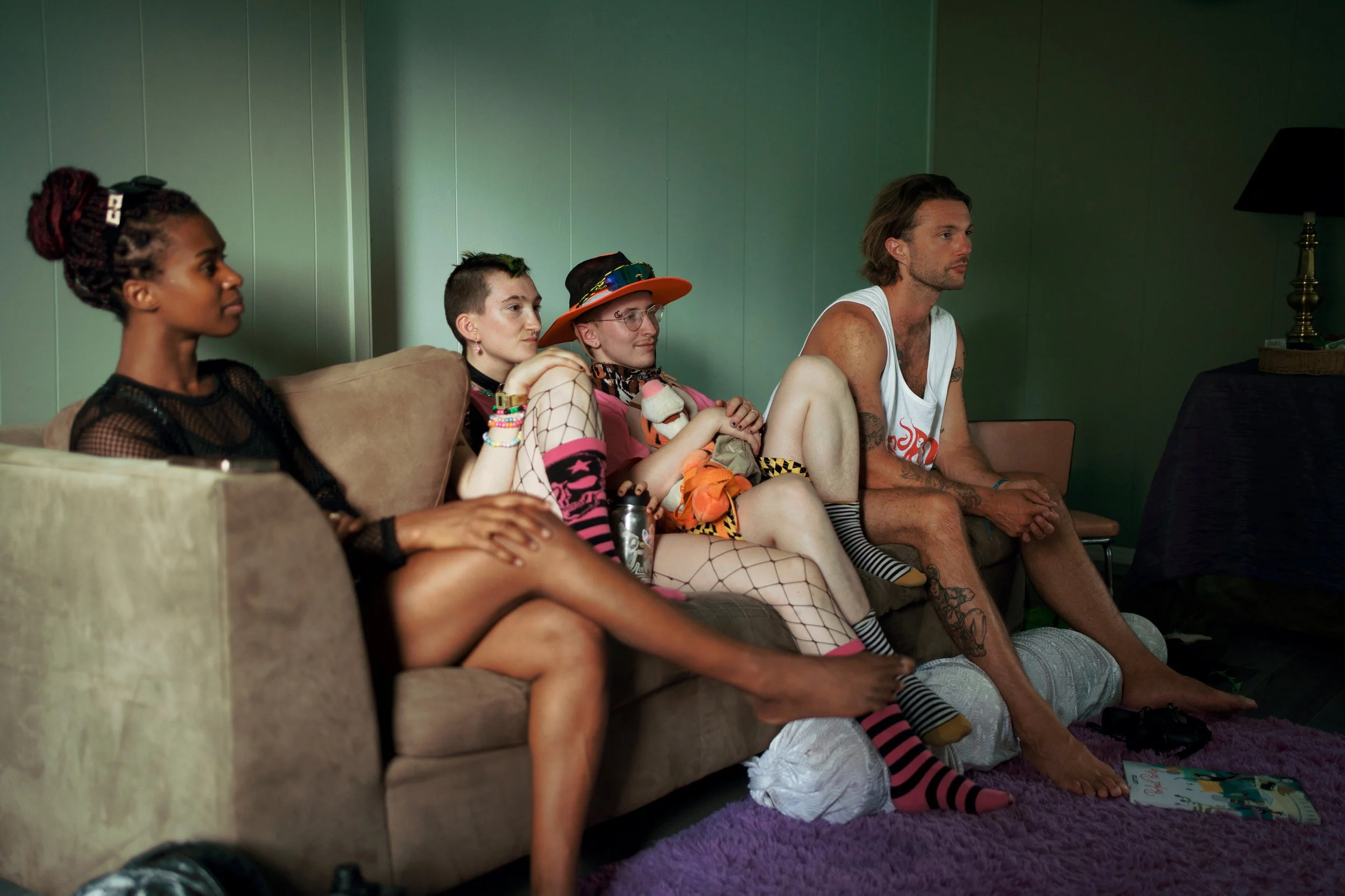 Four people sitting on a sofa watching TV in a room with green walls. The first person on the left is a woman with braided hair, wearing black clothing. The second person is a woman with short hair, with makeup and multiple bracelets, resting her chi