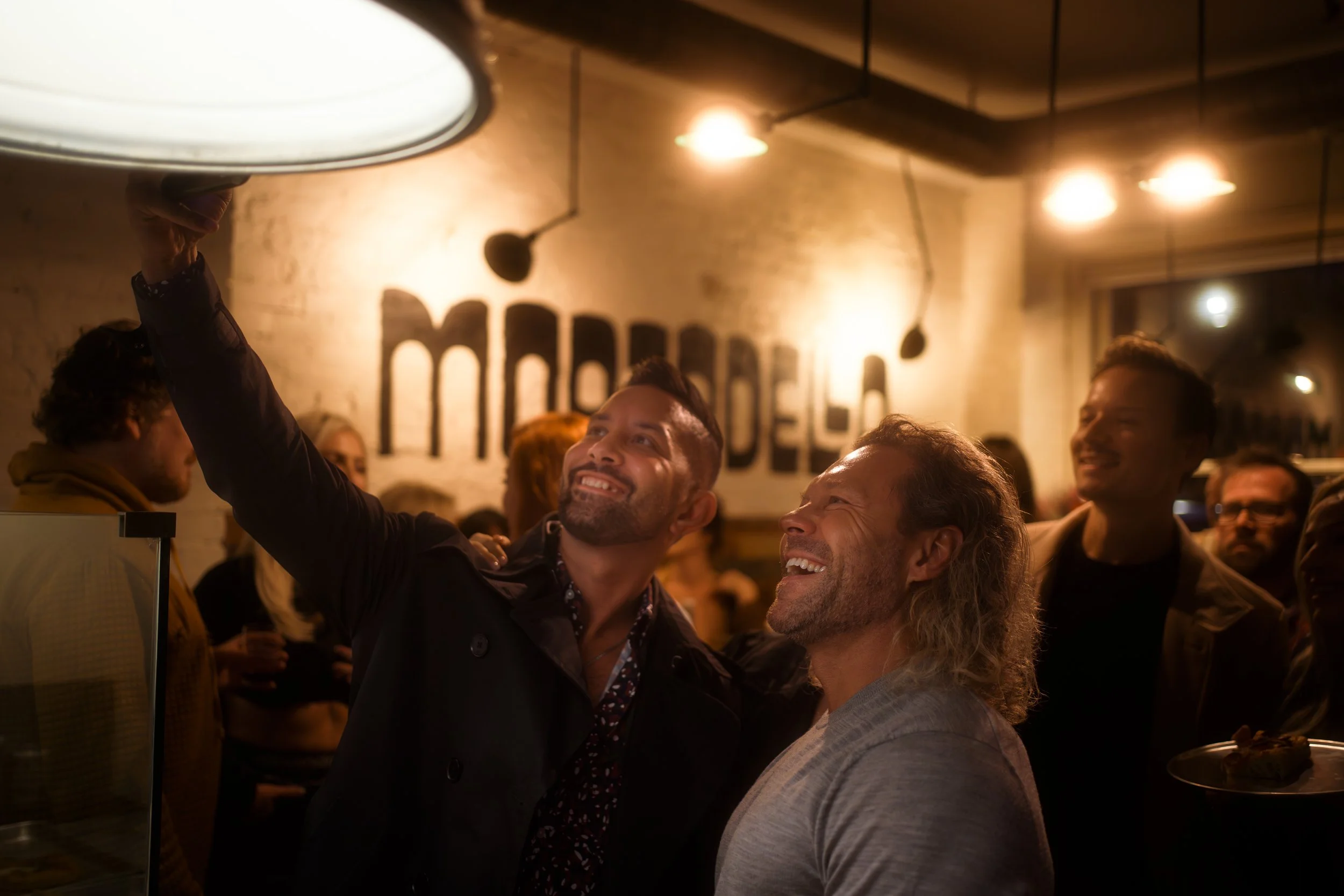 Two men taking a selfie together at a lively indoor gathering, smiling, with other people in the background and warm lighting.