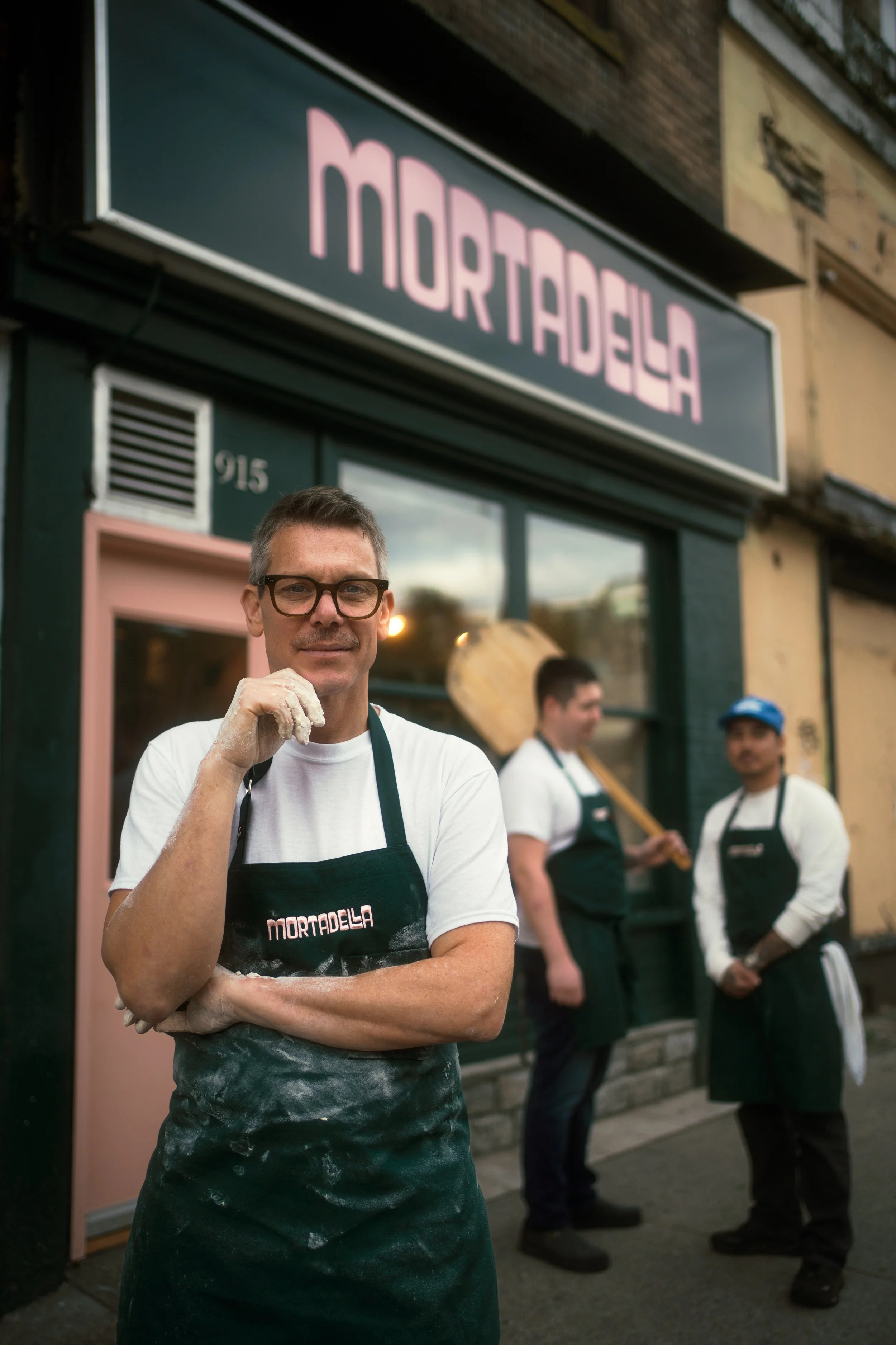 A man with glasses and a white T-shirt stands in front of a restaurant called Mortadela, wearing a black apron with the restaurant's name. Behind him, two men are talking outside the restaurant, one holding a large wooden spoon. The restaurant has a black and pink sign and a purple door.