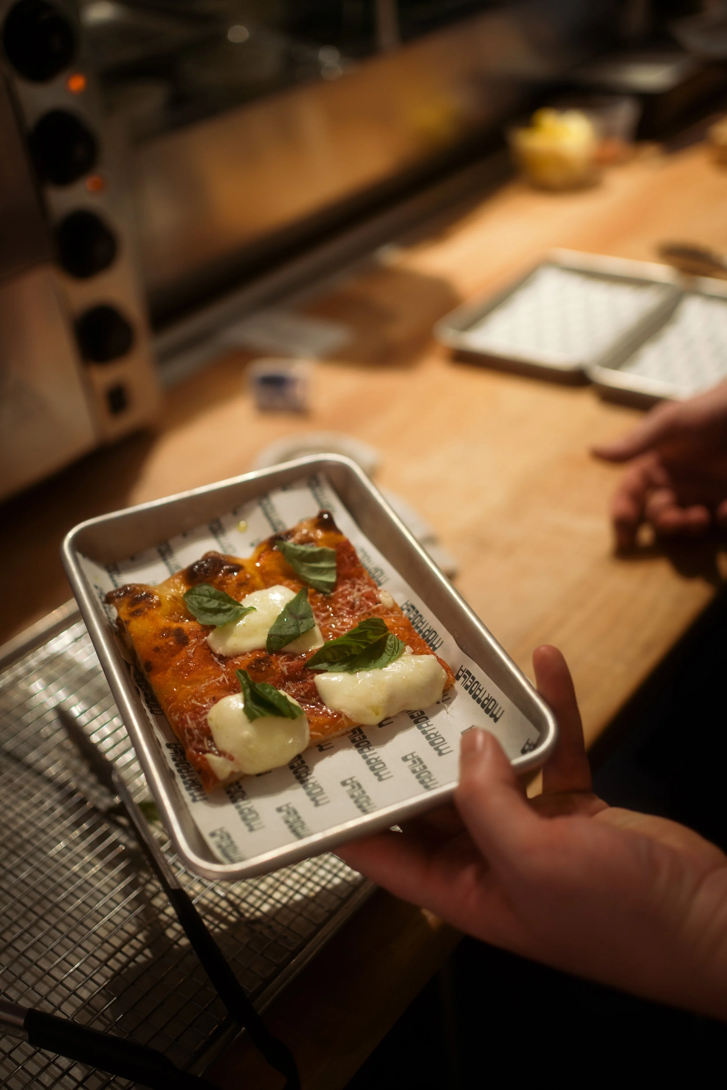 Person holding a tray with a slice of pizza topped with basil leaves and mozzarella cheese.