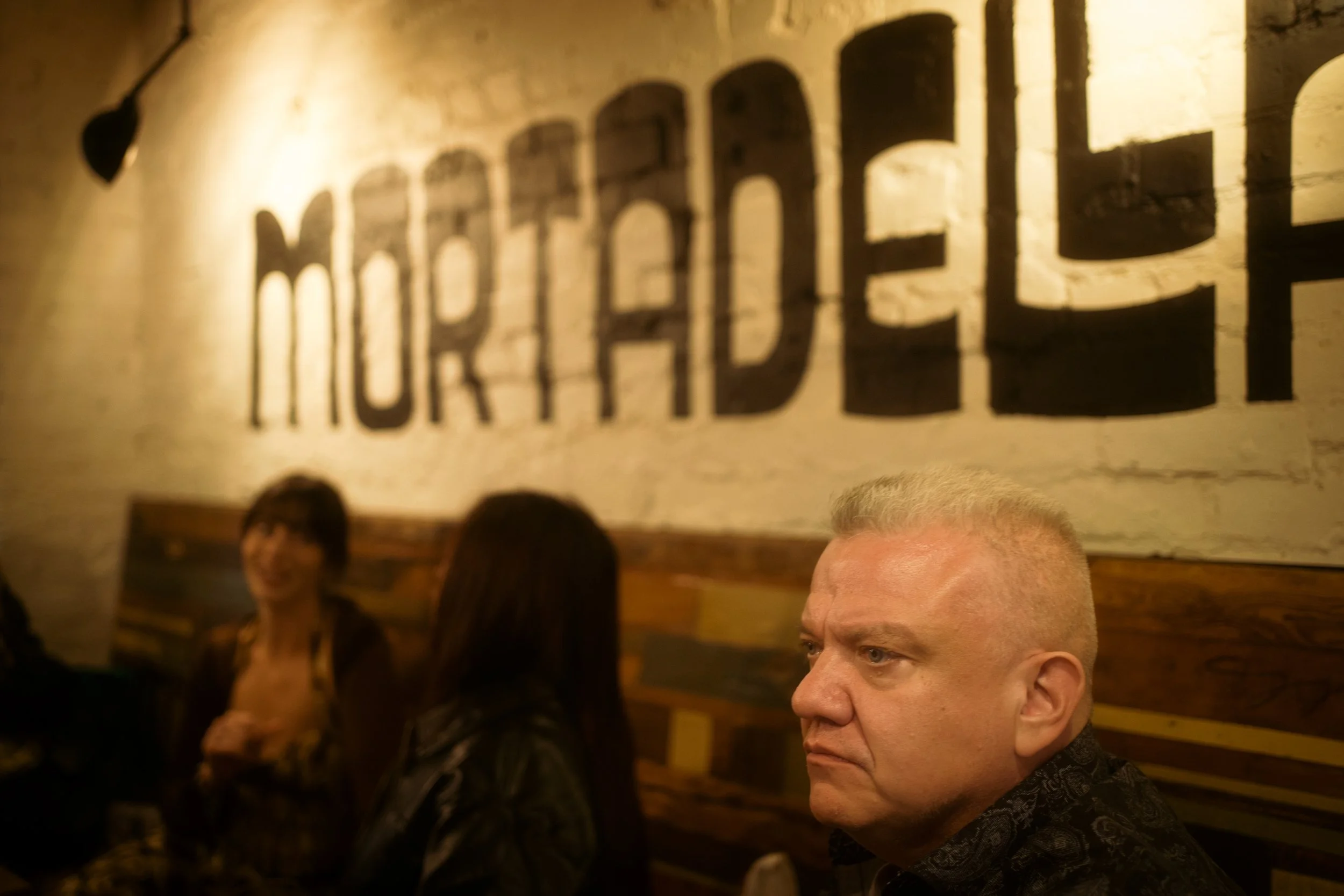 A man with short gray hair sits at a table in a restaurant or bar with a brown and beige color scheme. Behind him on the wall, the word 'MOREHEAD' is painted in large black letters on a white brick wall. Other people are sitting at the table, includi