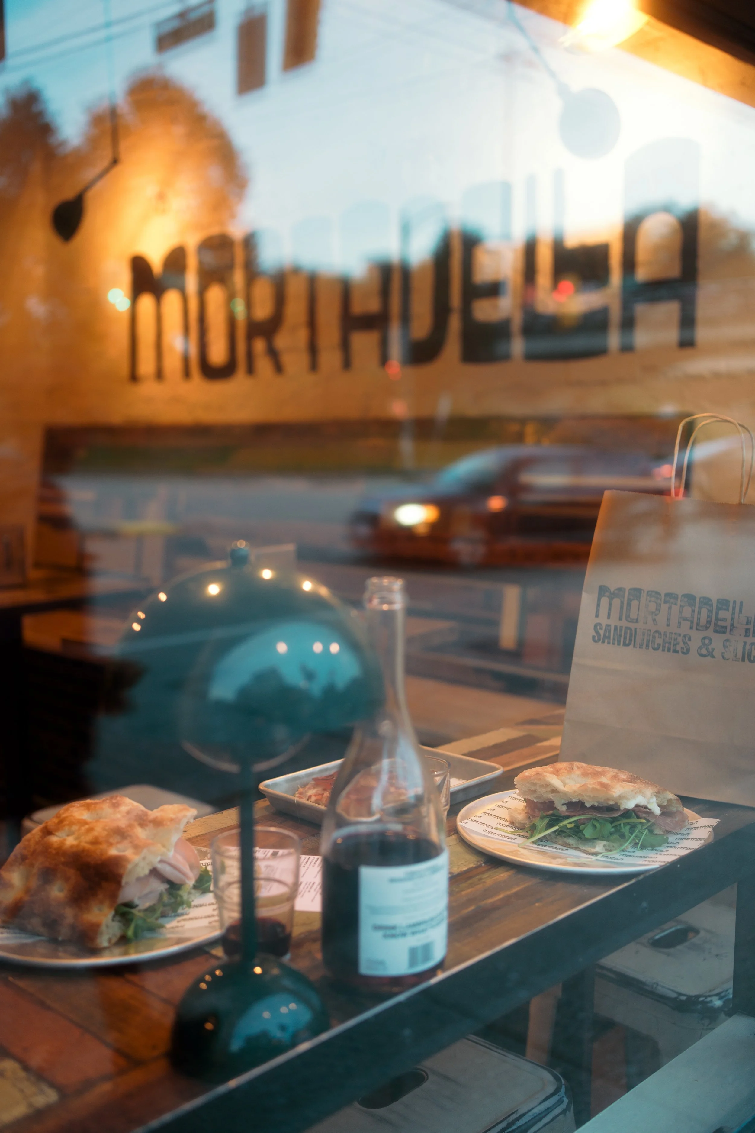 View through a restaurant window showing two sandwiches with greens on plates, a bottle of sauce, a small glass, and a small oven lamp, with the restaurant's sign partially visible and a blurred car passing by outside.
