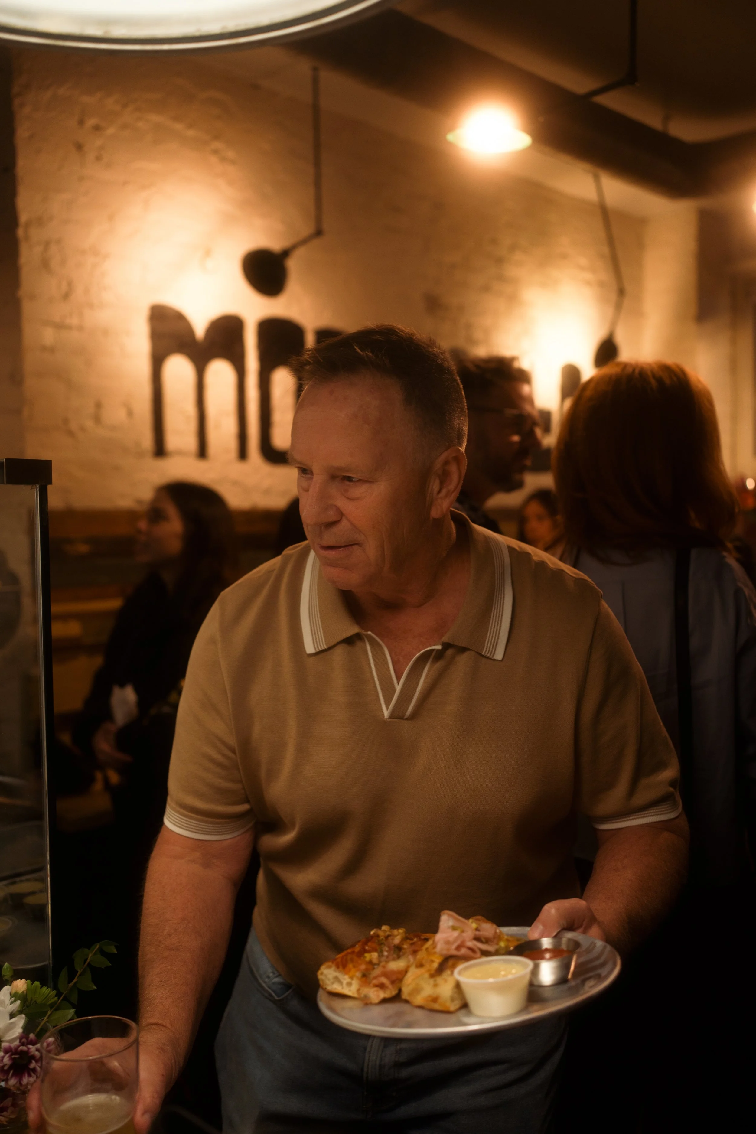 A man in a beige polo shirt holding a plate with pizza and dipping sauce in a restaurant with warm lighting and brick walls.