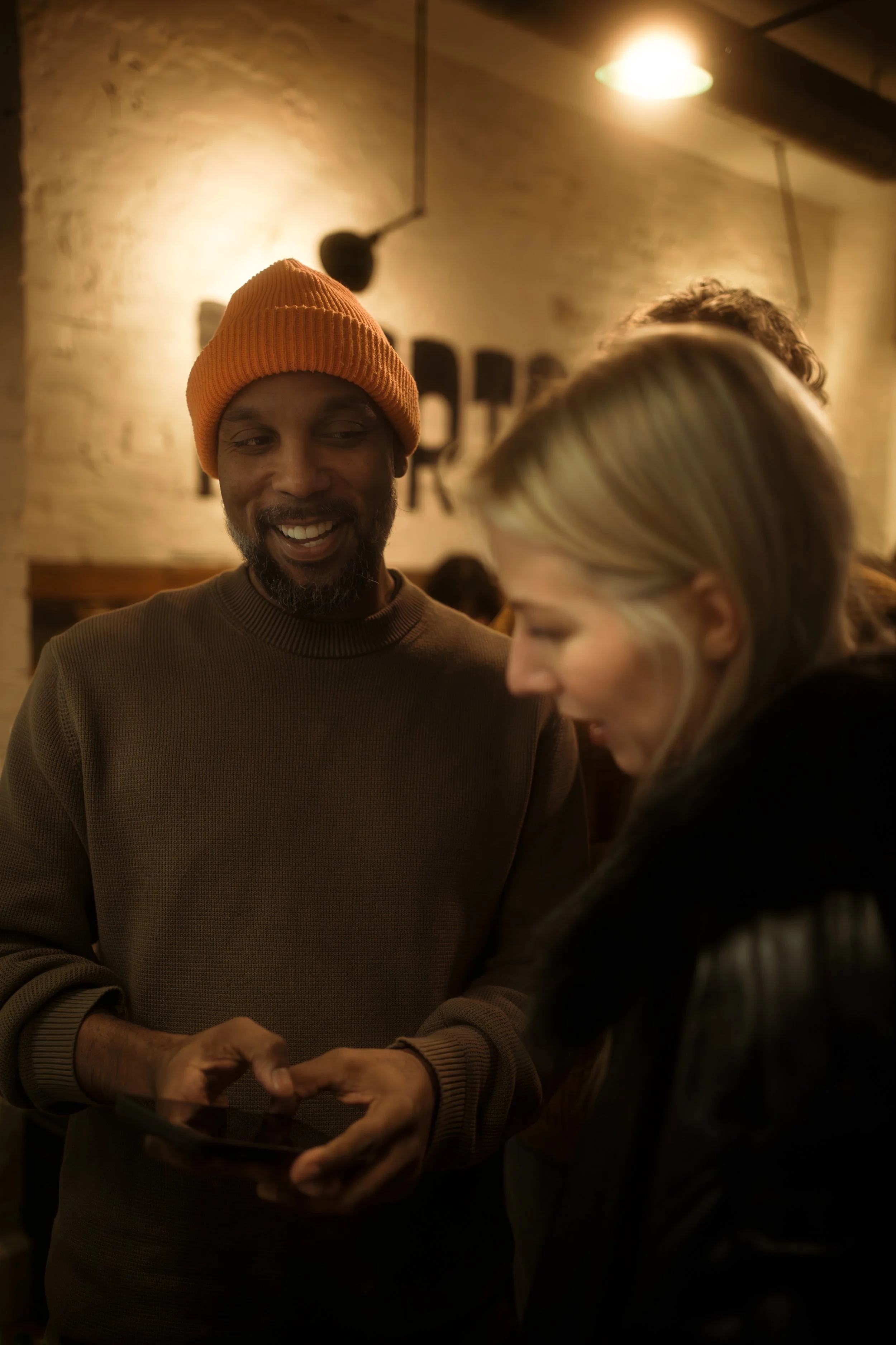 Two people smiling and looking at a phone in a warmly lit indoor setting, likely a bar or restaurant.