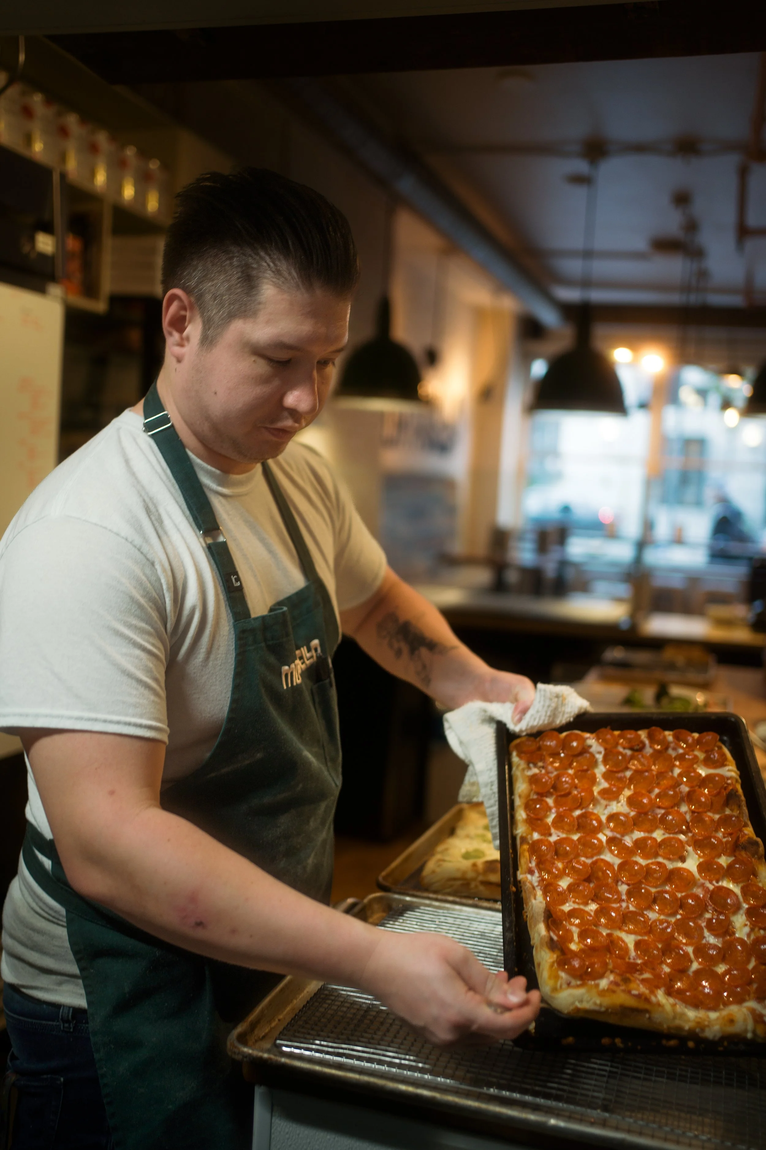 A man wearing a white t-shirt and a green apron is holding a large rectangular pizza with pepperoni slices in a restaurant kitchen.