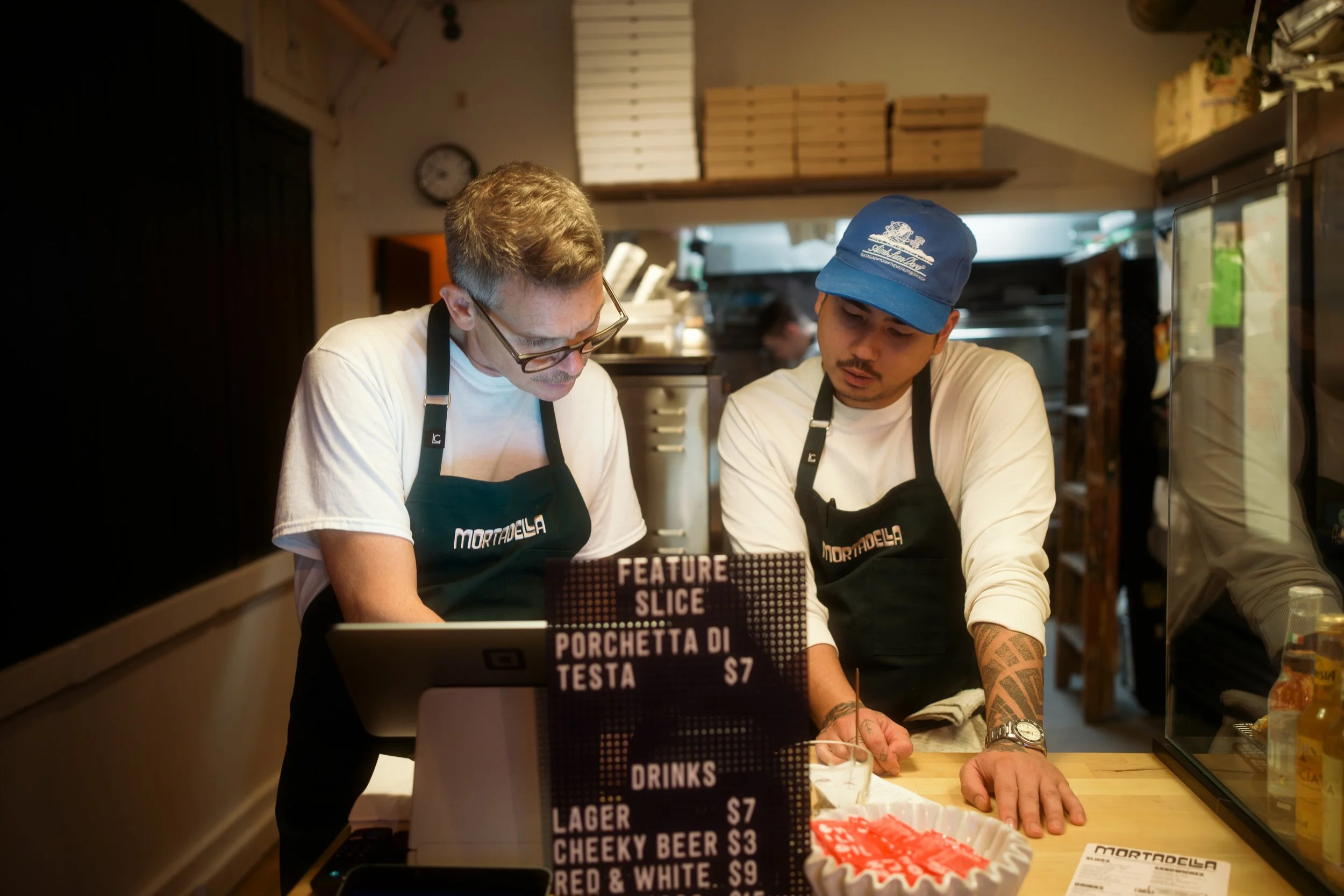 Two men working behind a restaurant counter, one with glasses and a mustache, the other wearing a blue cap, both wearing black aprons, discussing an order with a sign displaying menu items in front of them.