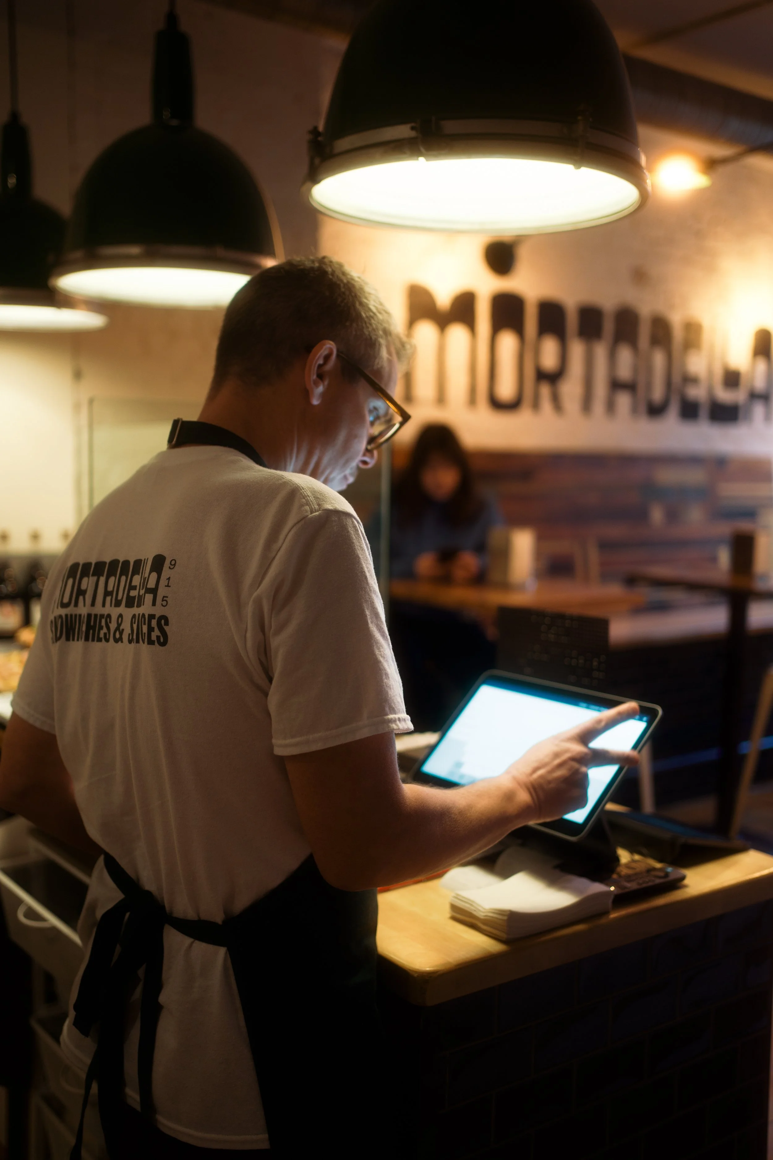 A man in a white T-shirt with a black apron, standing at a wooden counter, using a tablet in a dimly lit restaurant or cafe with hanging lights and a sign on the wall in the background.