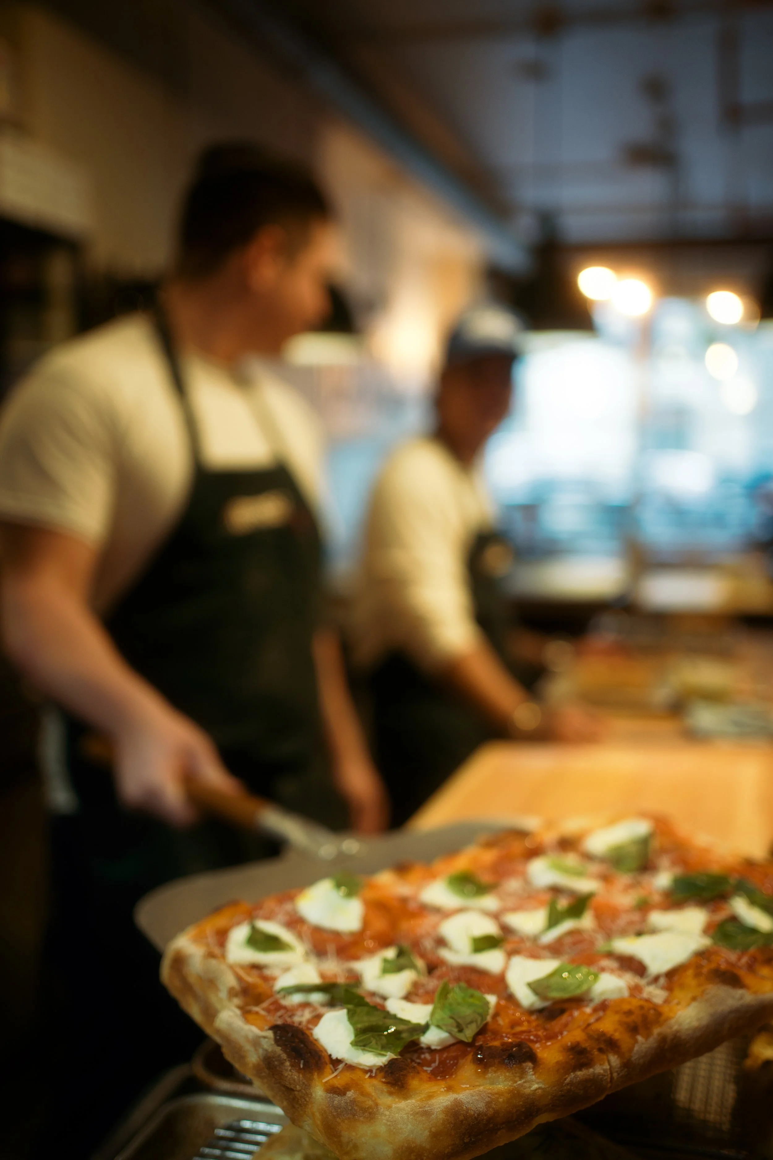 A slice of pizza with cheese, basil, and tomato toppings in the foreground, two chefs in black aprons preparing food in a rustic kitchen in the background.