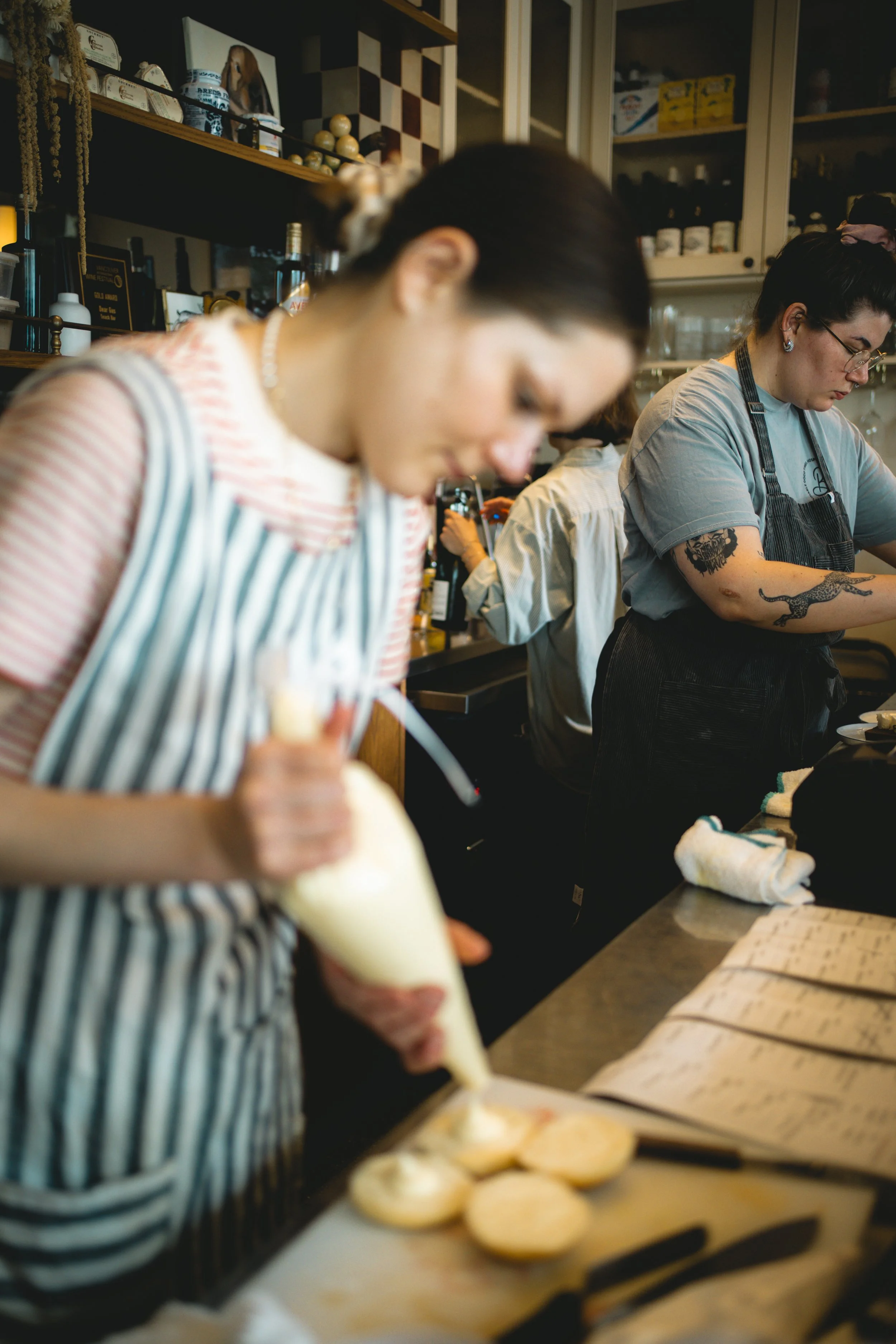 A woman in a striped apron decorating baked goods with icing in a busy kitchen or bakery.