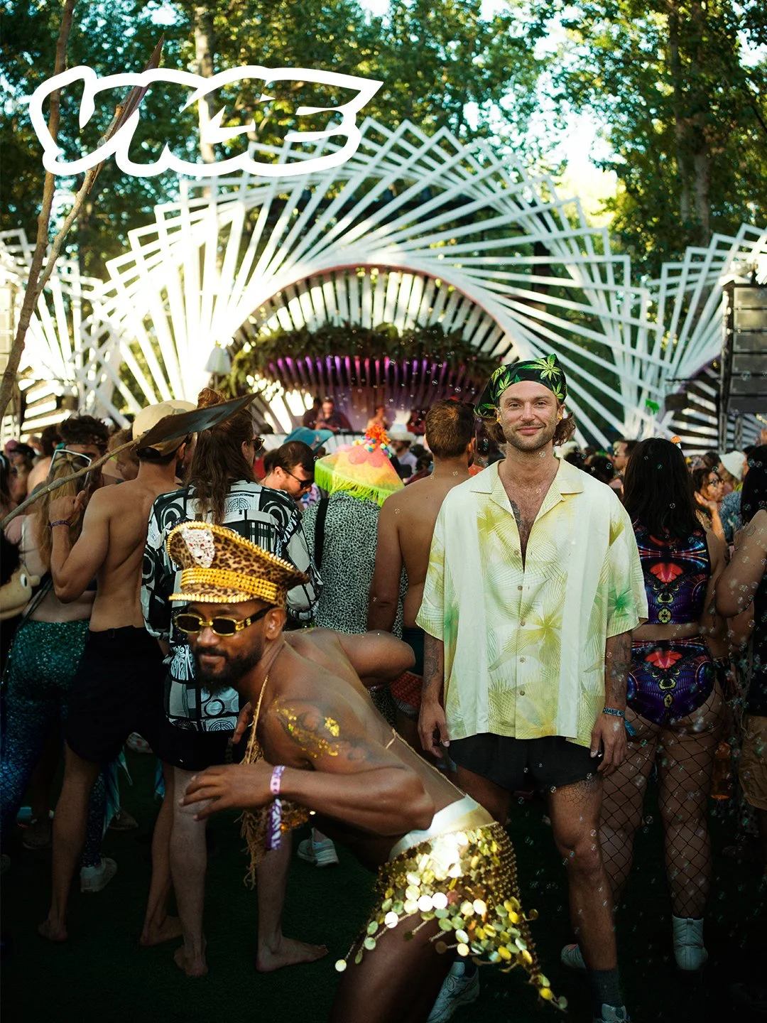 Crowd at an outdoor music festival with stage, trees in background, and colorful decorations. Two men in the foreground are dressed festively; one in gold sequins and the other in casual summer attire.
