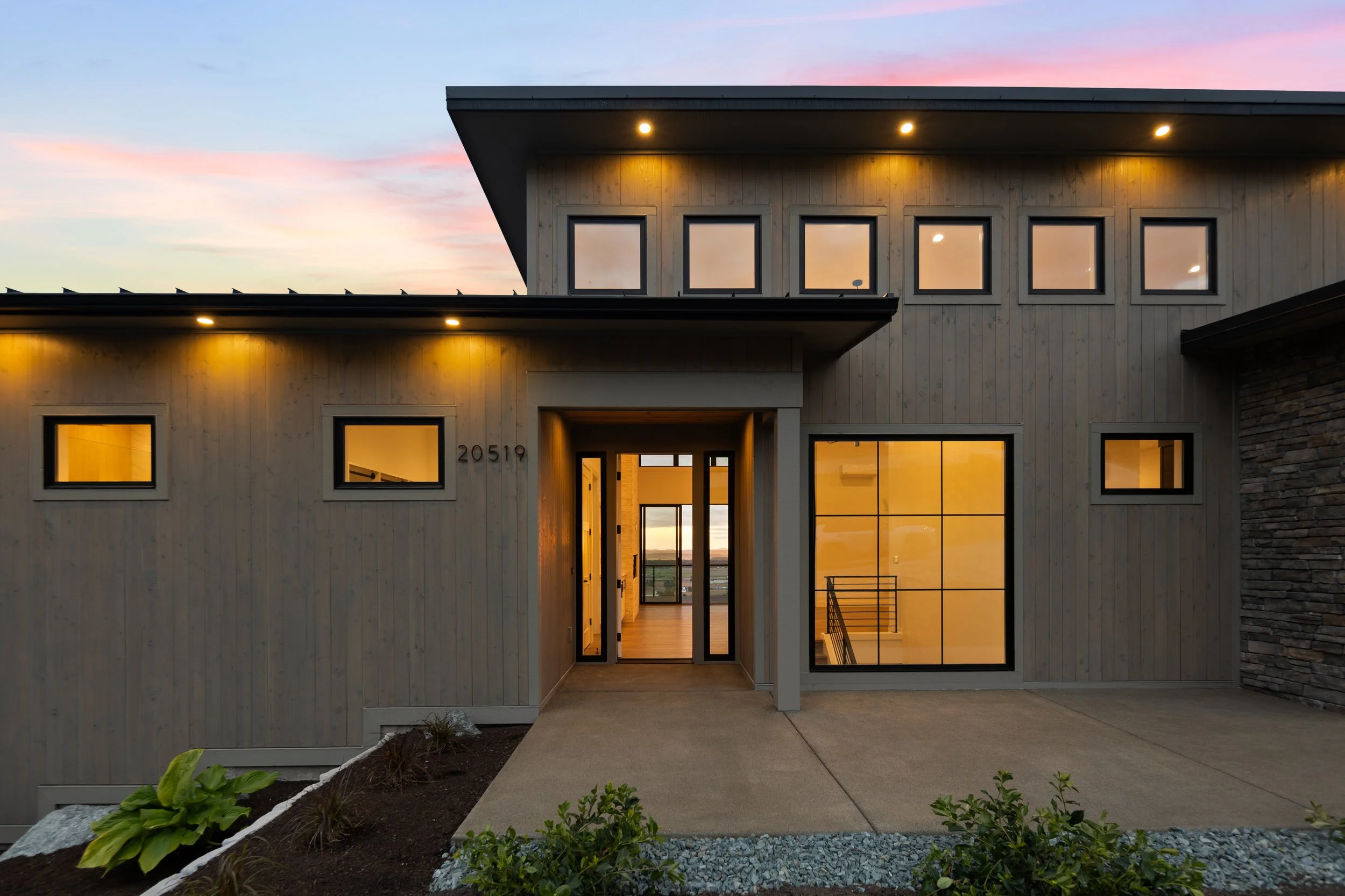 Modern two-story house with a beige wooden exterior, illuminated at sunset, with large windows and a glass entry door.