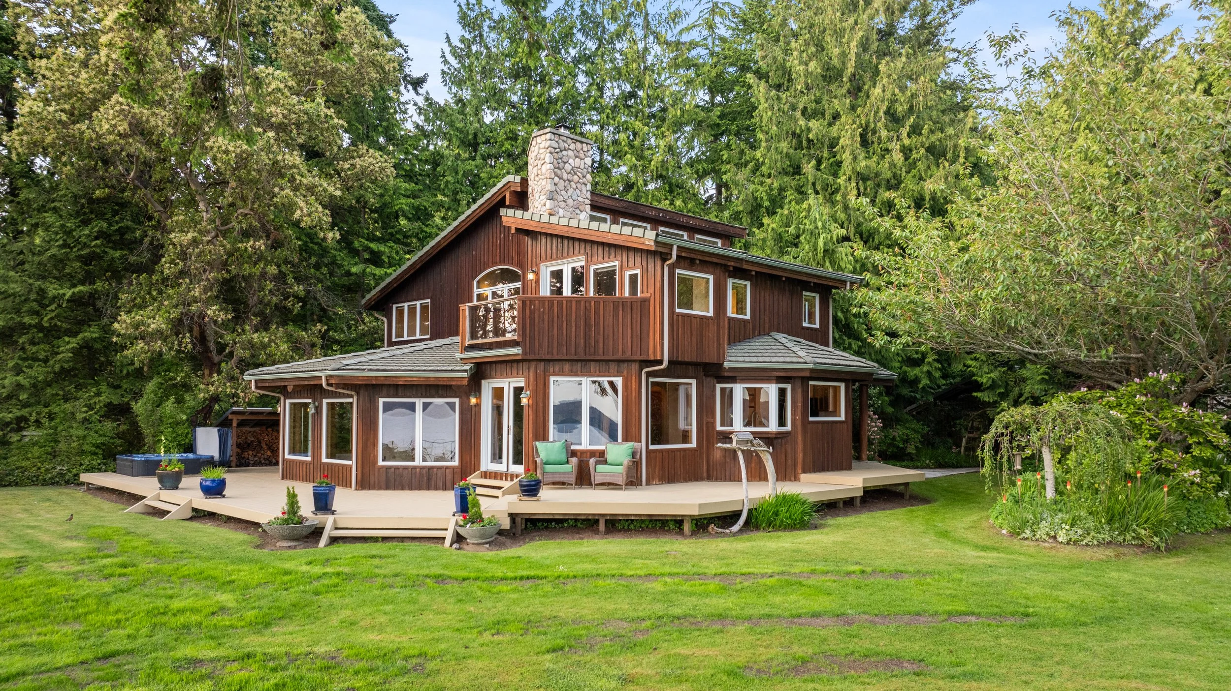 A modern two-story wooden house surrounded by green trees and a well-maintained lawn. The house has multiple windows, a chimney, and a spacious deck with outdoor furniture and potted plants.
