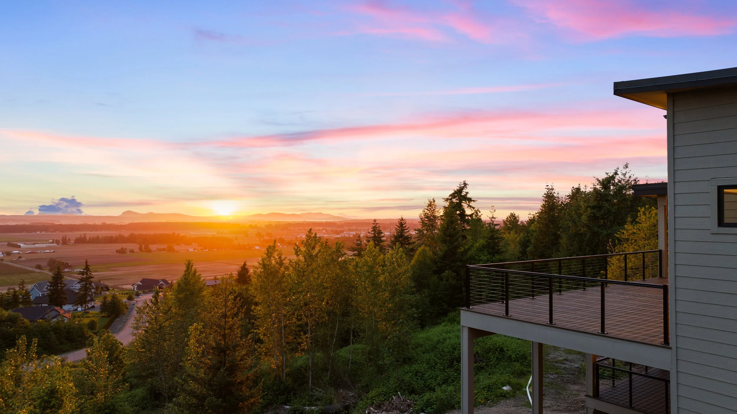 Sunset view over the countryside from a balcony, with trees in the foreground and a colorful sky with pink and purple clouds.