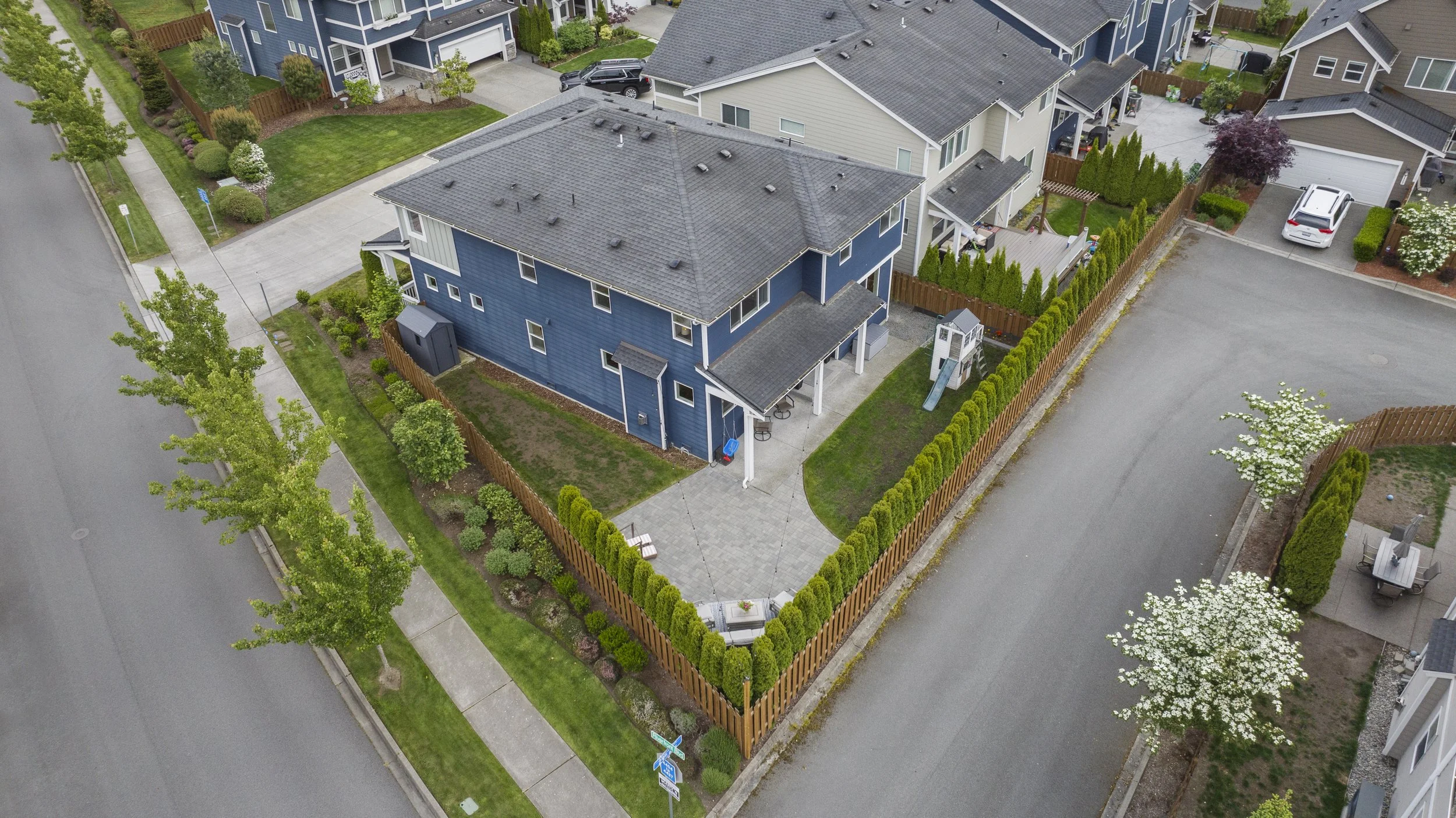 Aerial view of a blue two-story house with a fenced backyard, neighboring houses, trees, sidewalks, and a street.