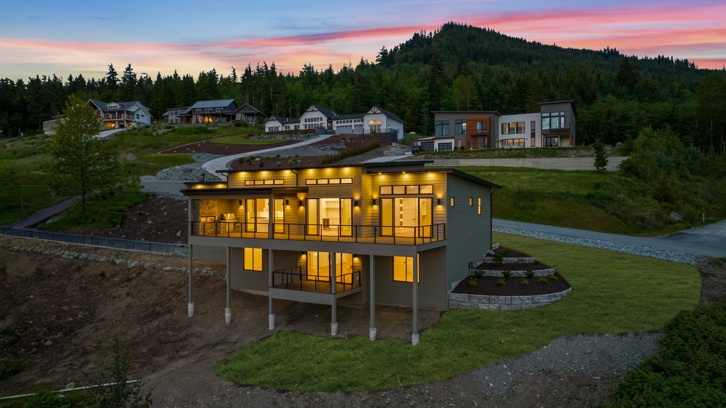 Hillside neighborhood houses at dusk, illuminated windows, modern architecture, surrounded by trees and greenery.