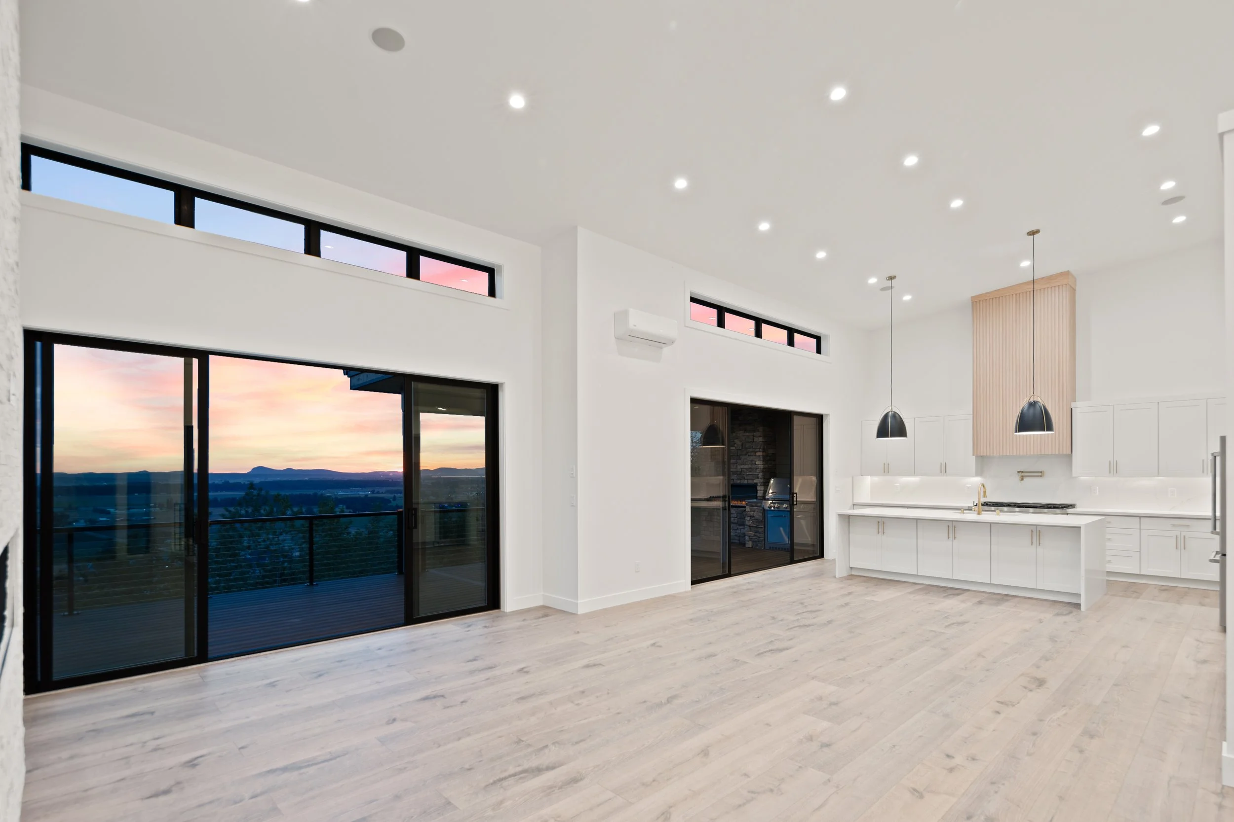 Modern open-concept kitchen and living space with large sliding glass doors leading to a balcony, overlooking a sunset landscape. The room features white cabinets, a kitchen island, black pendant lights, and high windows near the ceiling, with light wooden flooring and recessed ceiling lights.