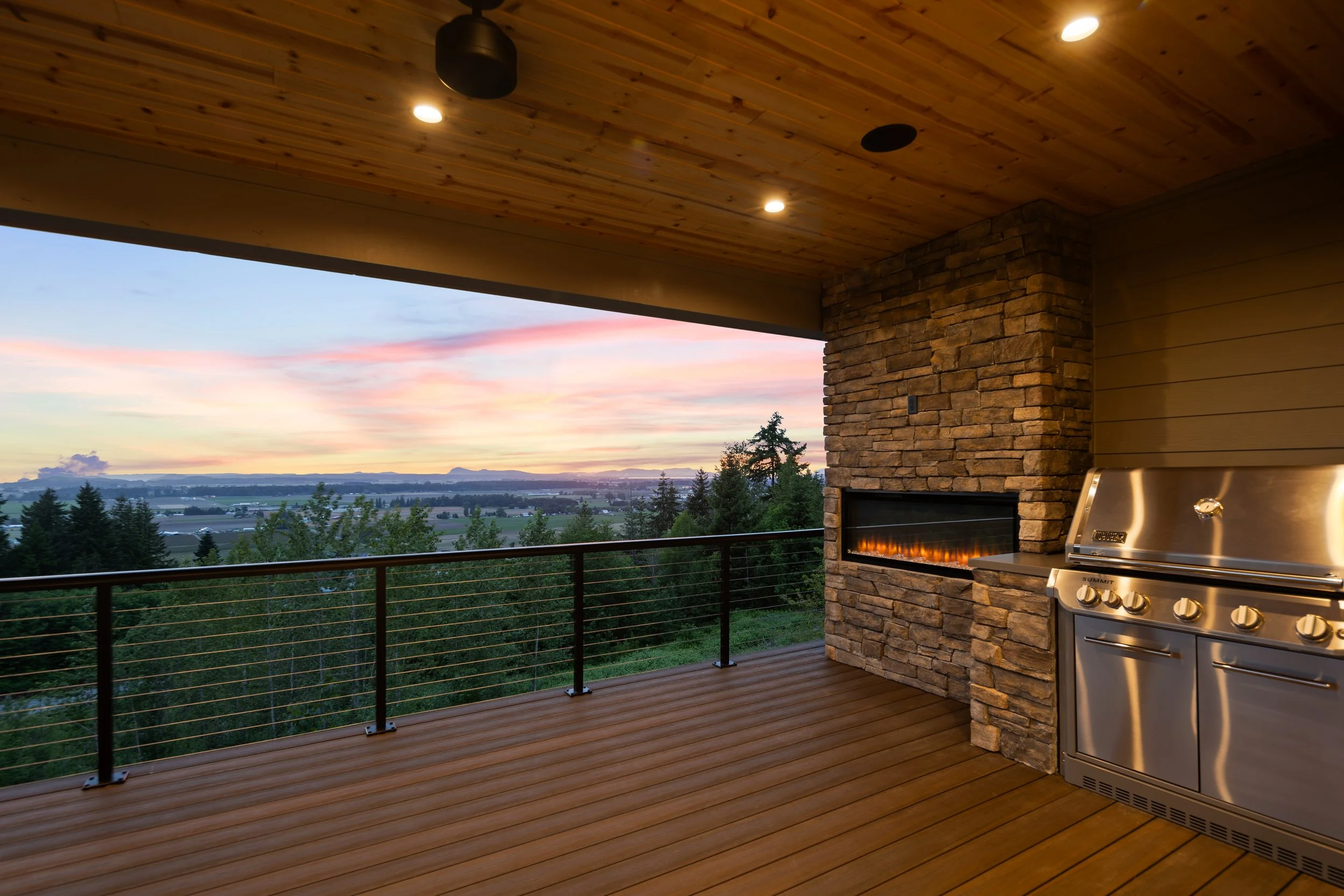 Outdoor balcony with a stone fireplace, stainless steel grill, wooden ceiling, and a view of trees and a colorful sky at sunset.