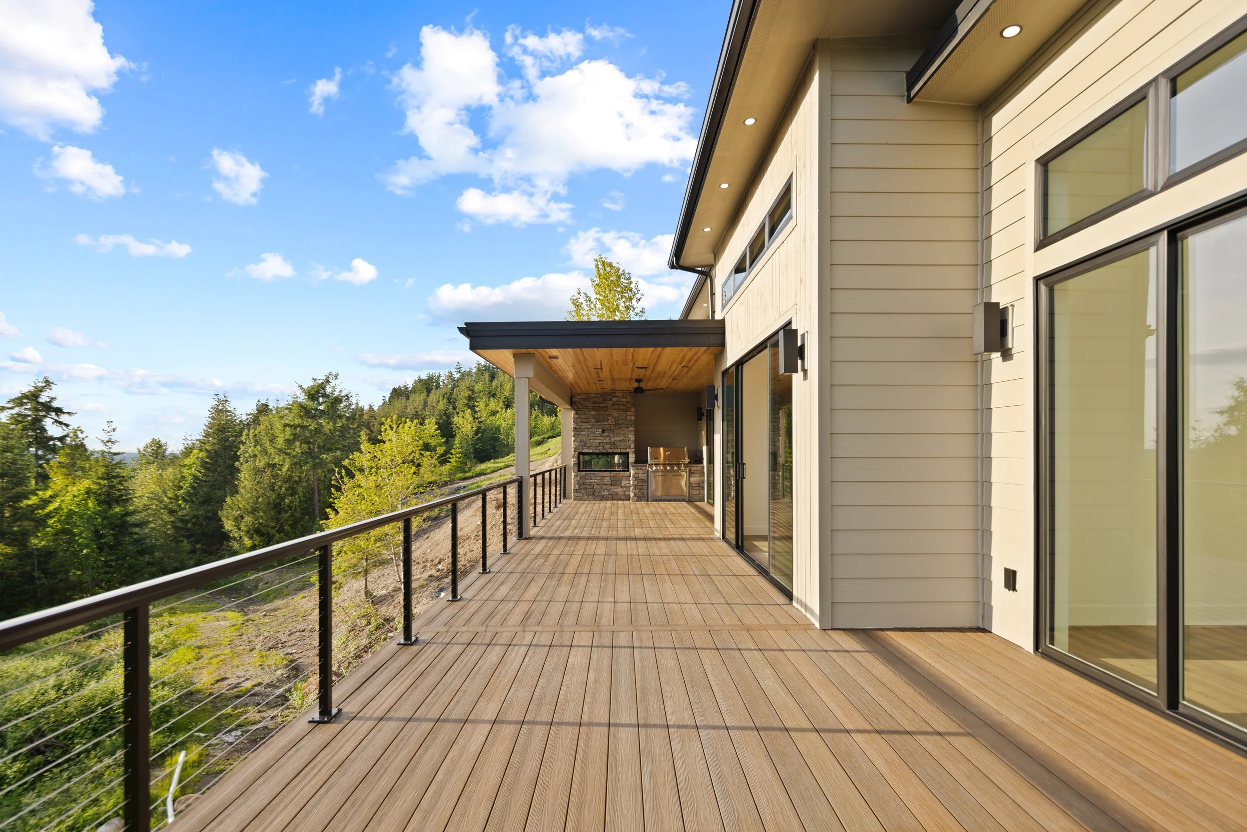 Modern outdoor balcony with wooden flooring, glass sliding doors, and a black metal railing, overlooking a lush green forest under a blue sky with scattered clouds.