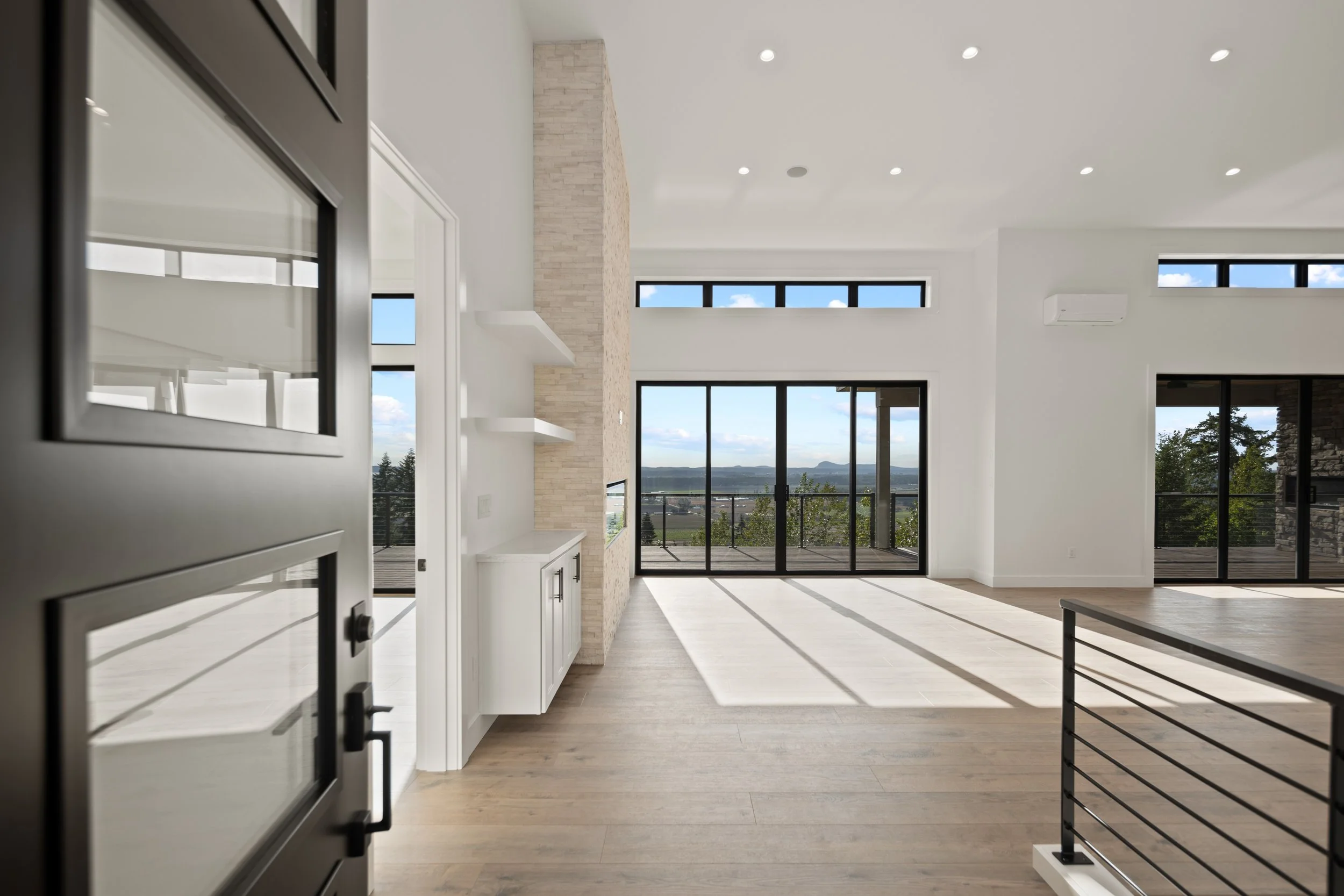 Interior of a modern, empty living room with large windows and sliding glass doors, light-colored wooden floors, white walls, and a balcony overlooking a scenic landscape.
