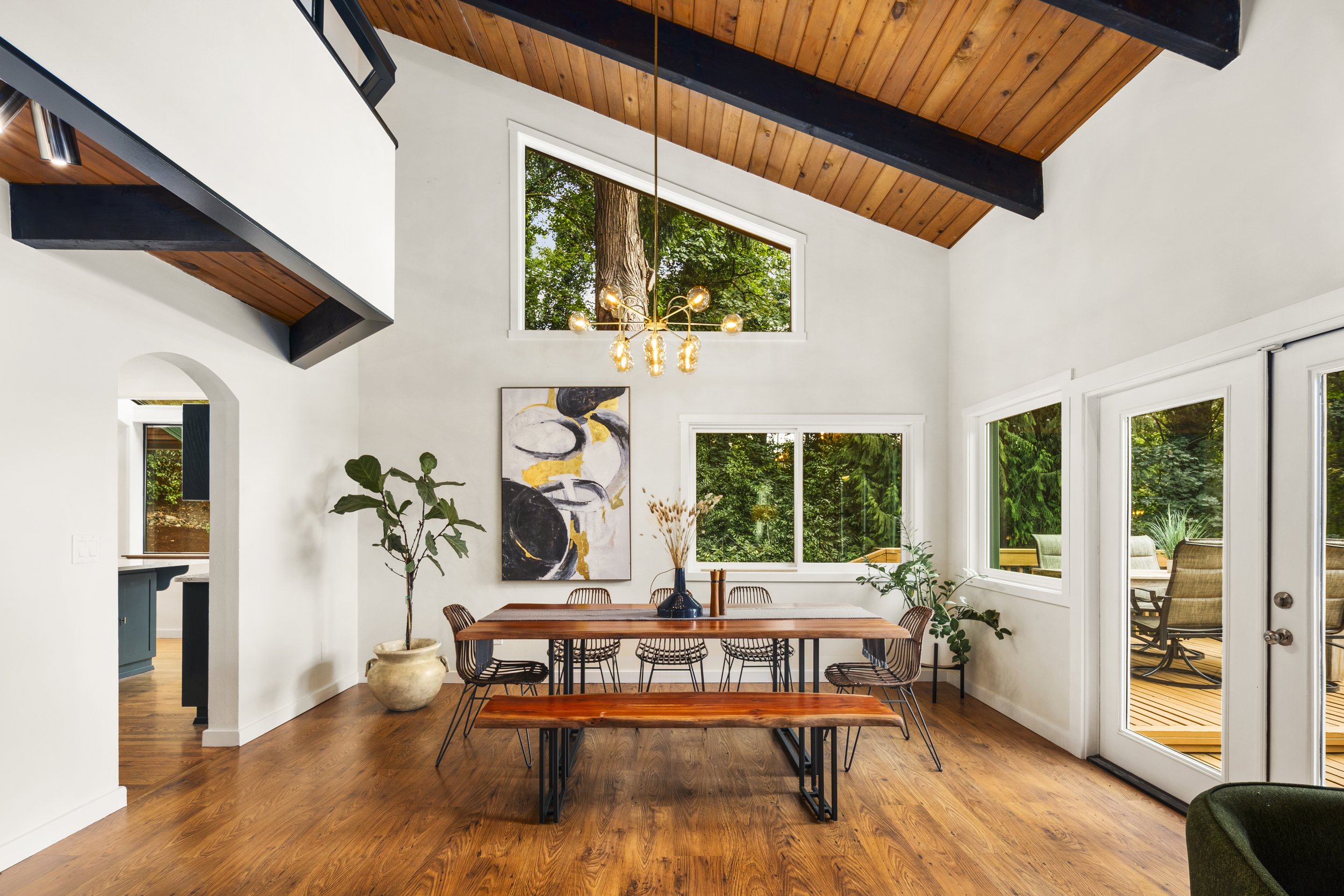 Modern dining room with wooden floor and white walls, featuring a wooden dining table with six chairs, large windows, and a sliding glass door leading to an outdoor patio. A chandelier hangs from the vaulted, wood-paneled ceiling, and there is artwork and potted plants for decor.