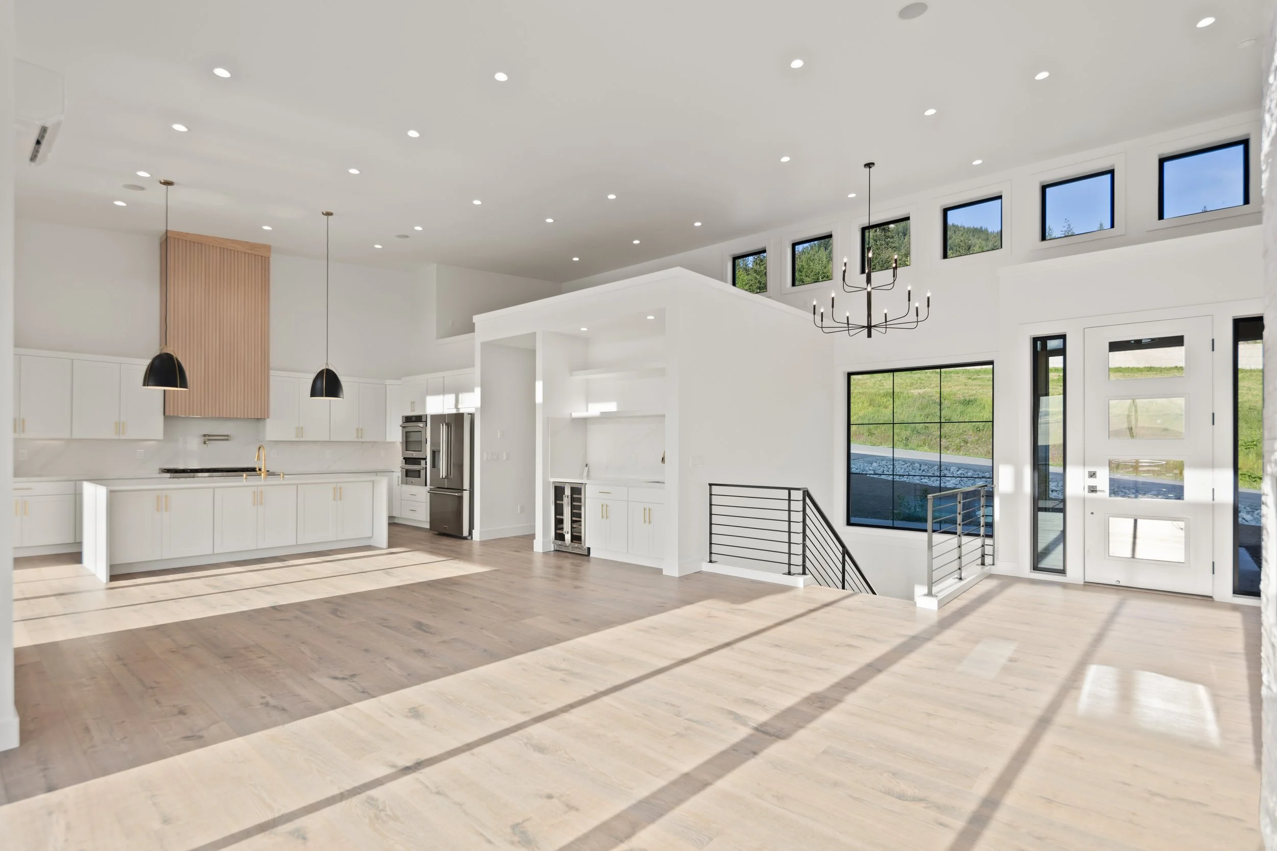 Empty living room and kitchen with white walls, large windows, wood flooring, and modern fixtures.