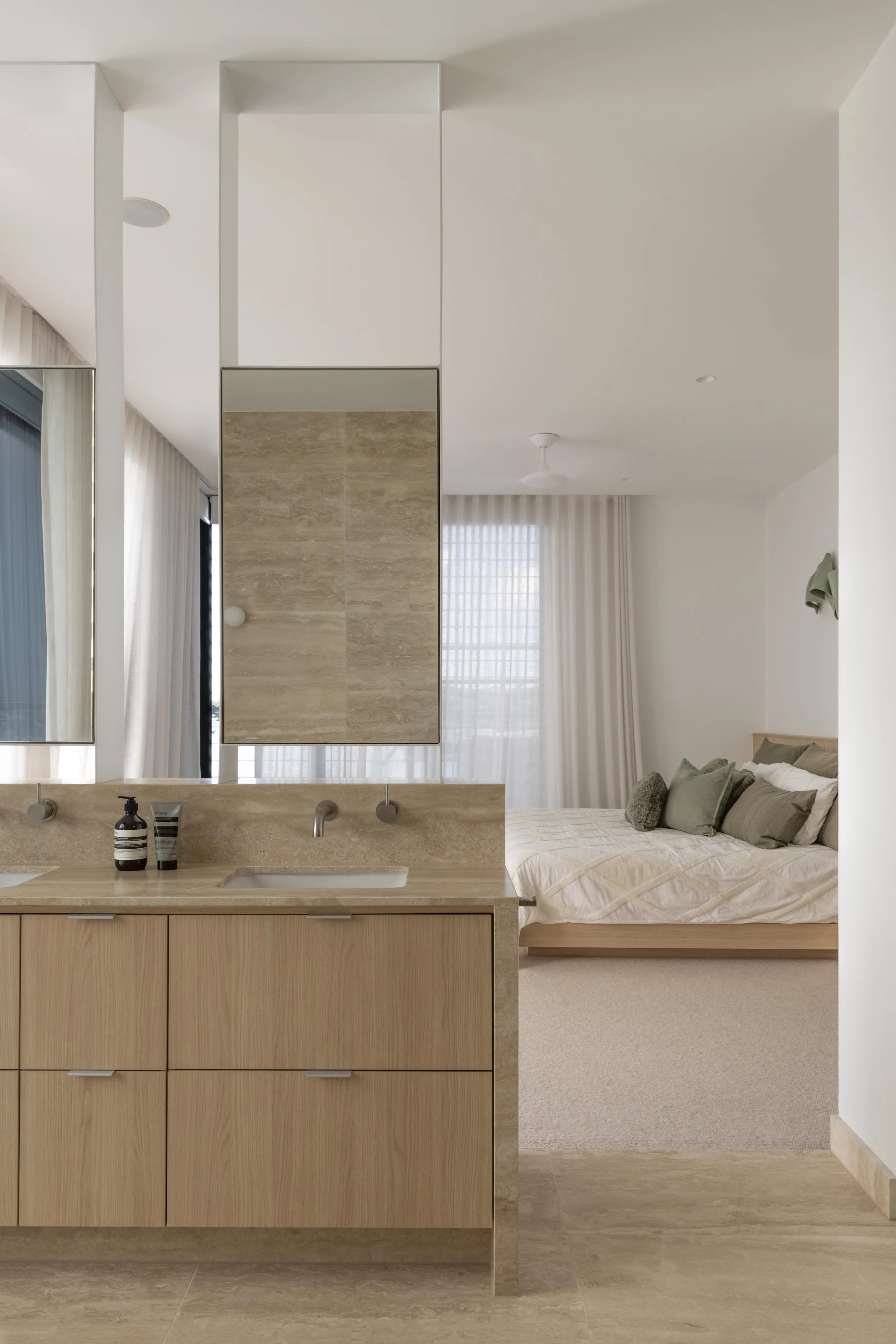 This refined ensuite at Project124 features a double stone vanity with warm timber cabinetry, vertical mirrors, and minimalist wall‑mounted tapware. The space flows seamlessly into the adjoining bedroom, where soft textiles, layered green and neutral