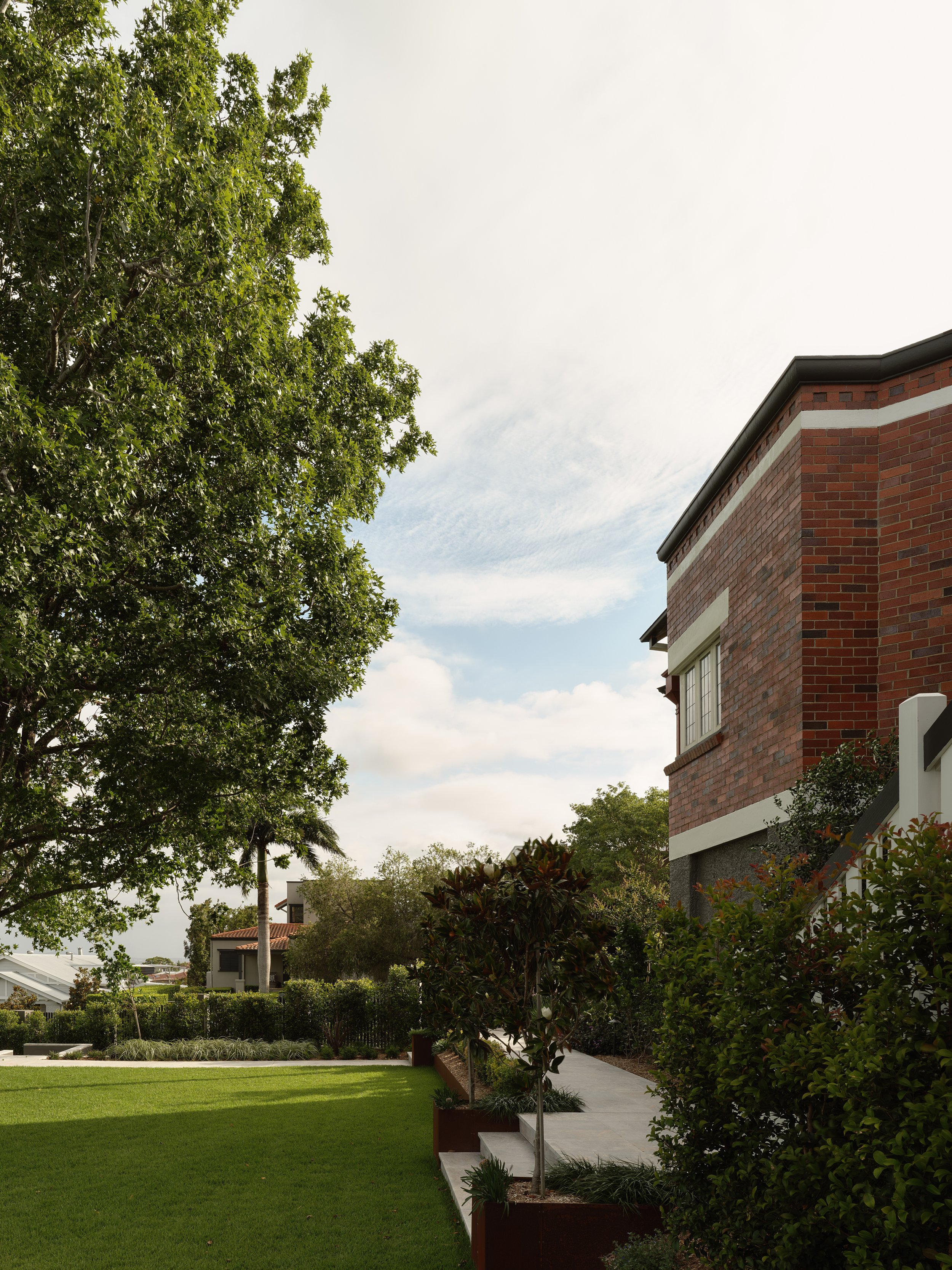 View of a residential backyard with a large tree, manicured lawn, bushes, and a brick house with stairs.