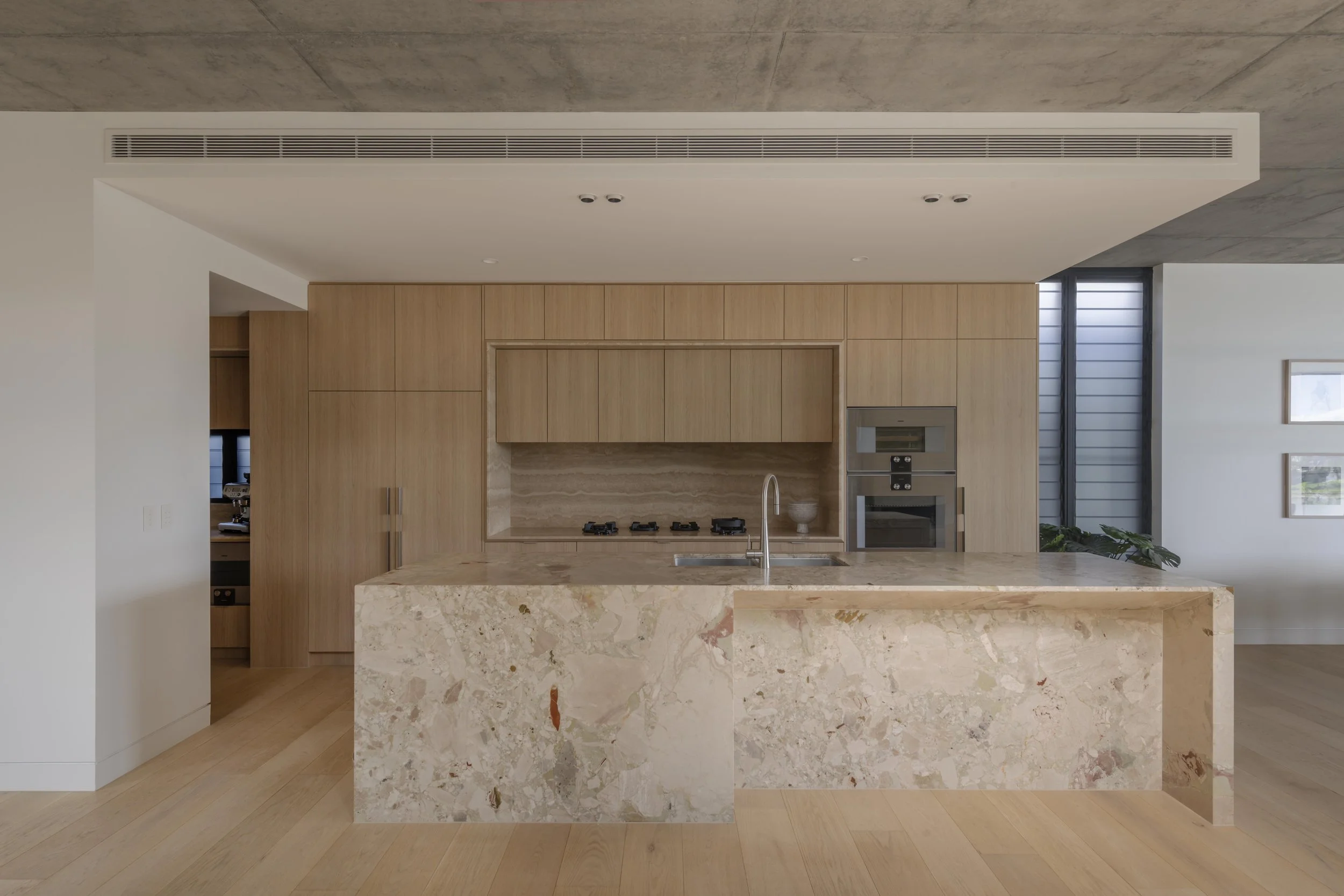 This contemporary kitchen at Project124 showcases a sculptural stone island with rich natural veining, warm timber cabinetry, and integrated appliances set within a clean, minimalist layout. Recessed lighting and a streamlined ceiling vent emphasise 