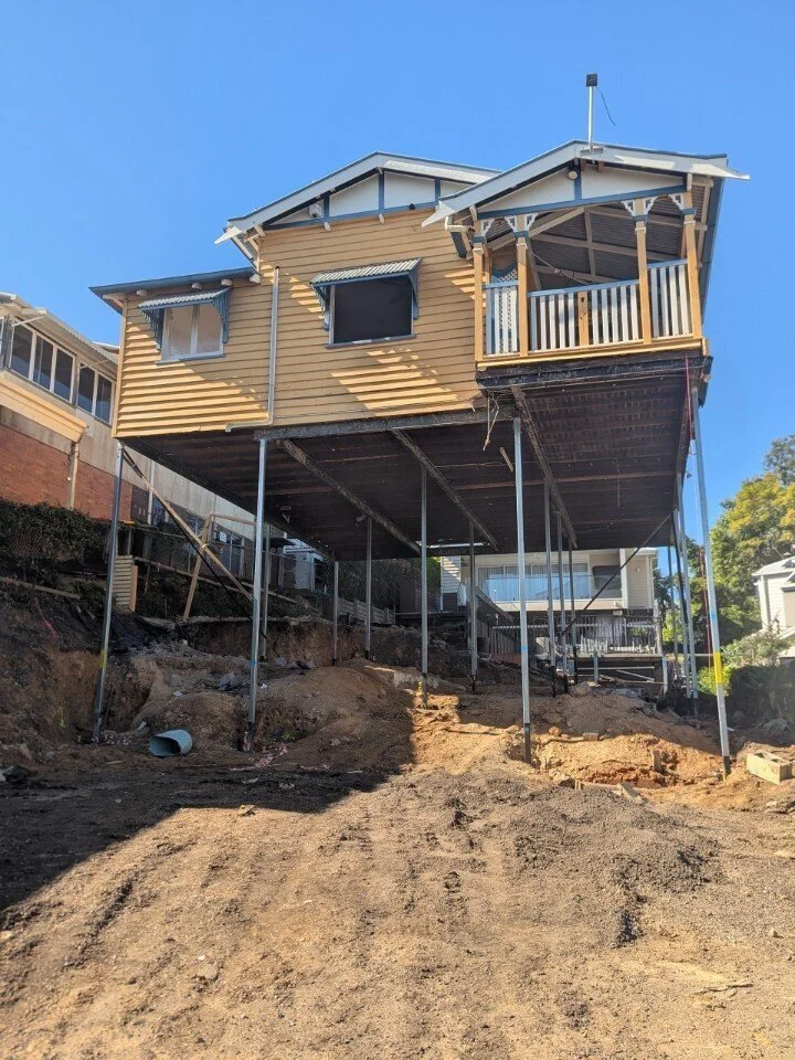 A timber house is elevated high on steel supports above an excavated construction site, with exposed soil, rocks, and service pipes visible beneath the structure. The surrounding homes sit on sloping ground, highlighting the complexity of the house‑r