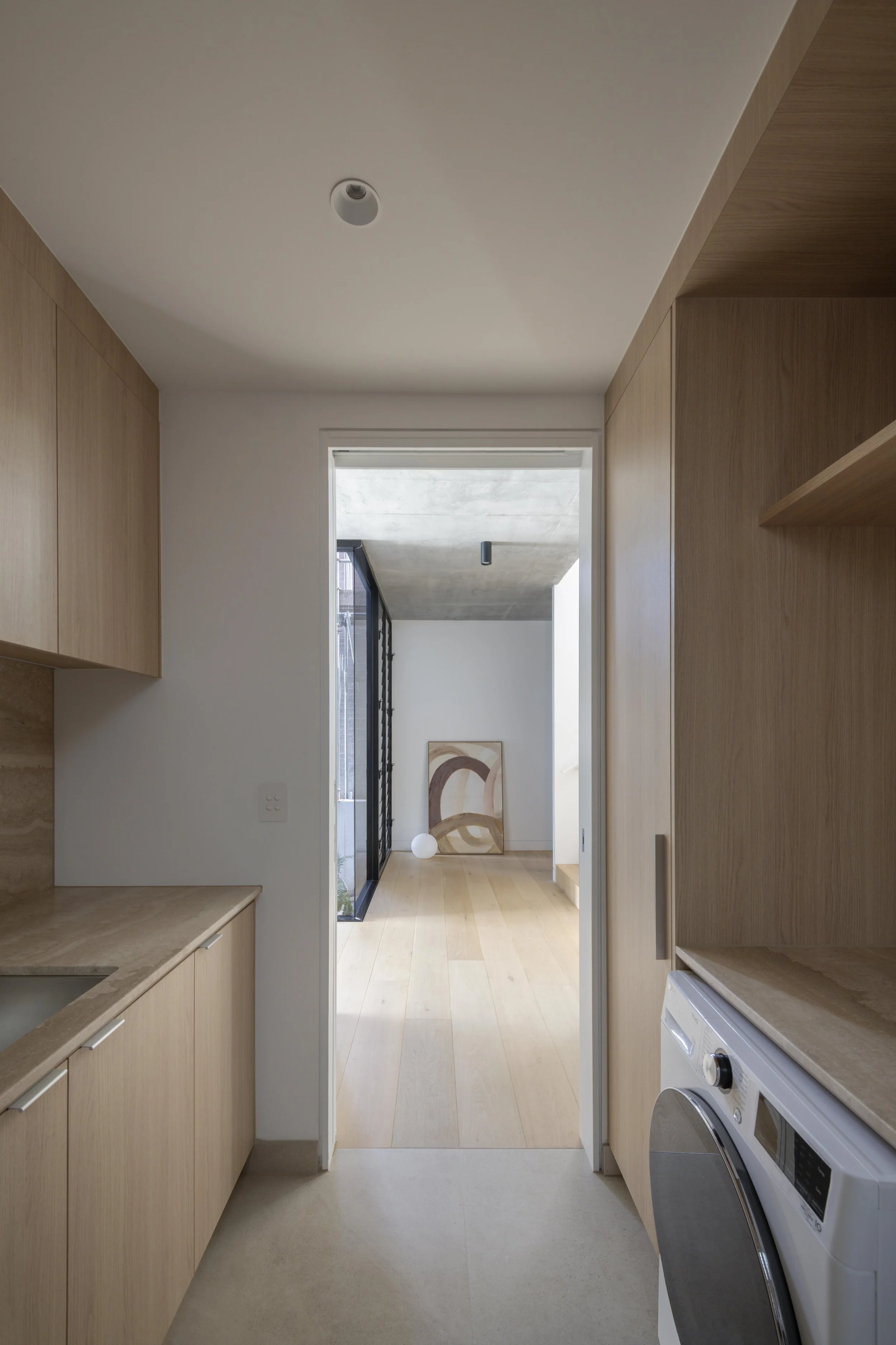 This refined utility space at Project124 features custom timber cabinetry, integrated appliances, and a clean transition from tiled flooring to warm timber in the adjoining room. Natural light fills the corridor beyond, where curated artwork and expo