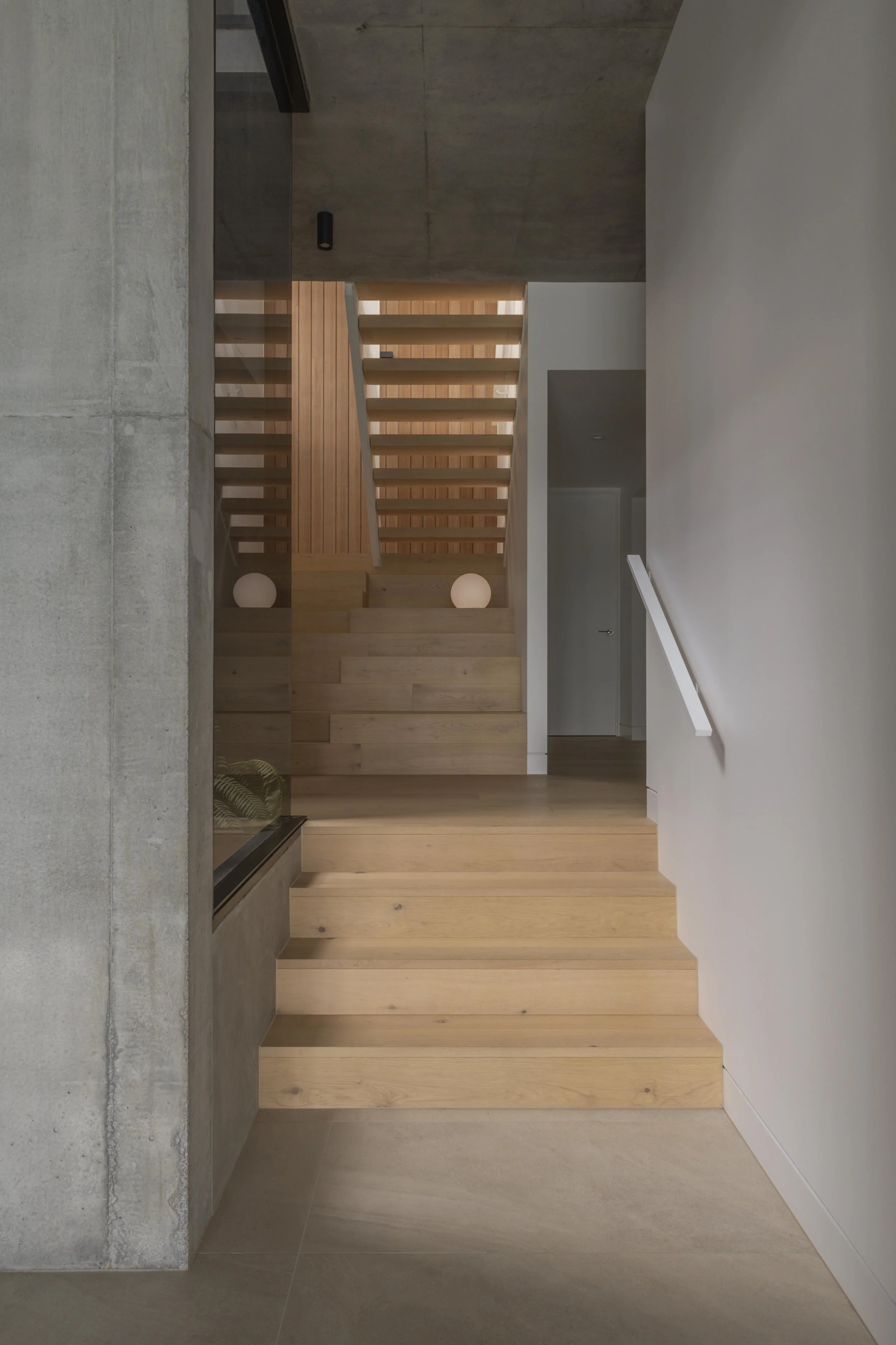 This sculptural timber staircase at Project124 features wide lower treads, open‑riser upper steps, and soft spherical lighting that highlights the warm material palette. Exposed concrete ceilings and crisp white walls frame the minimalist composition