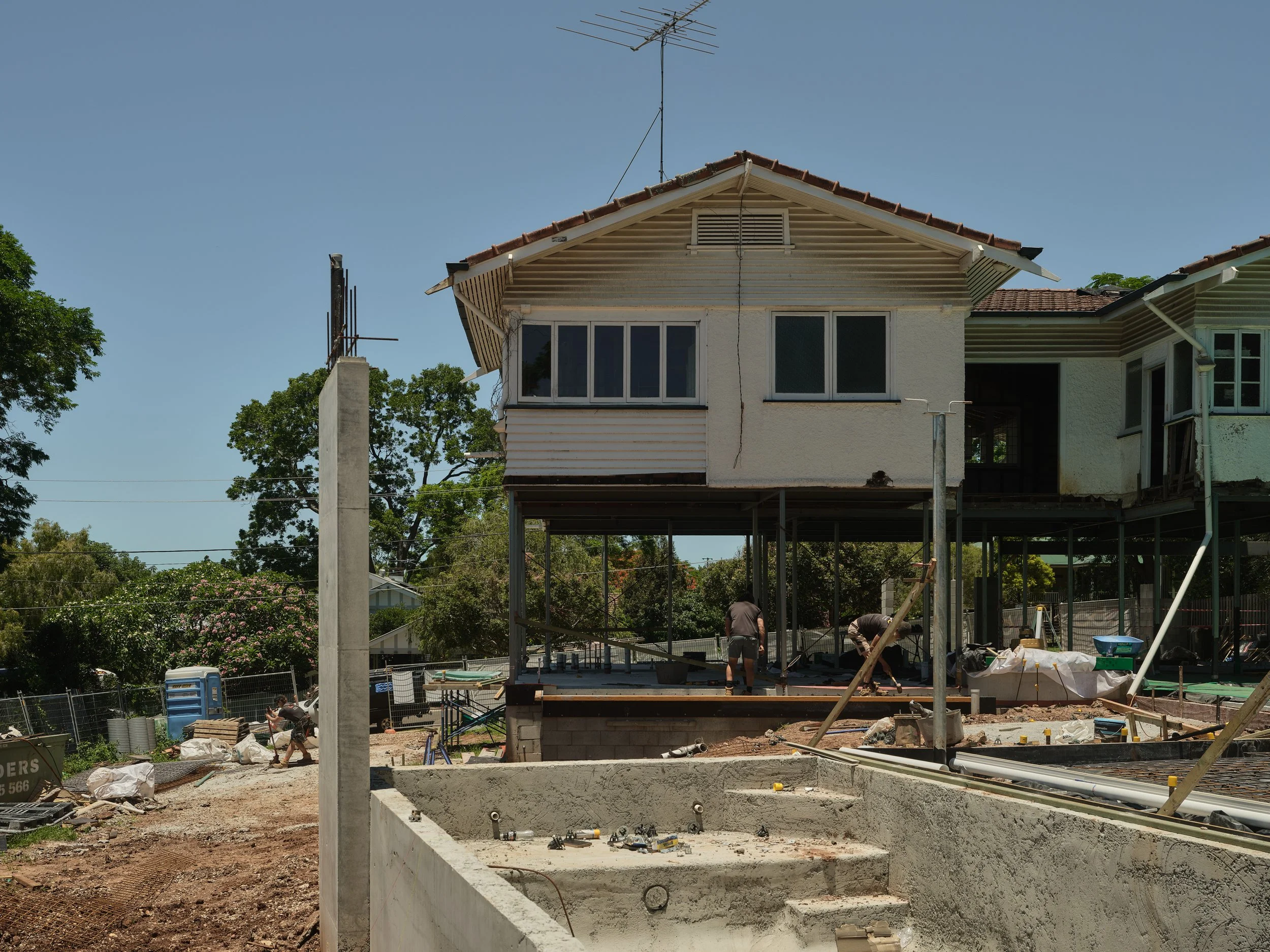 Major architectural renovation in progress at Project 134, showing the existing home elevated on structural supports while the new lower‑level build‑under and pool area take shape. Construction crews are onsite completing structural works around the 