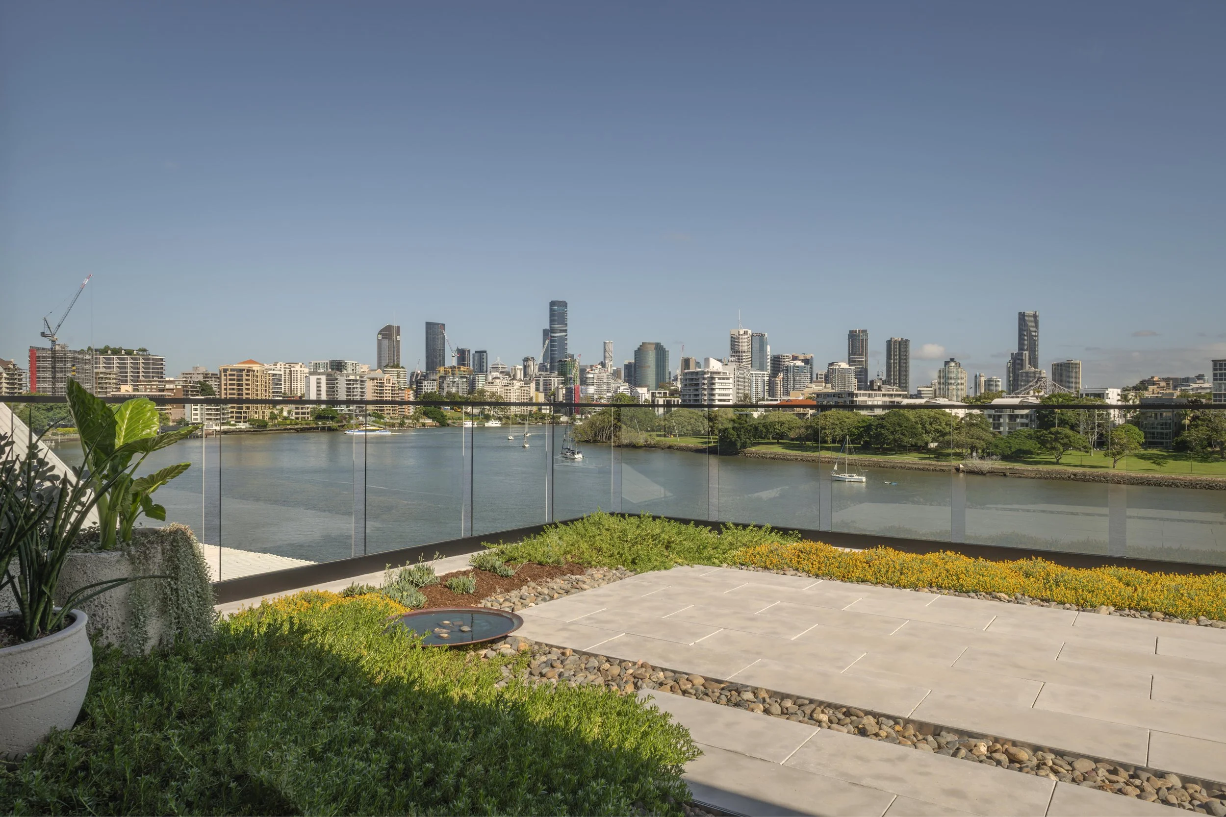 This elevated rooftop garden at Project124 overlooks sweeping river and city skyline views, blending lush planting, a sculptural water feature, and stone pathways within a calm, contemporary landscape design. A frameless glass balustrade preserves th